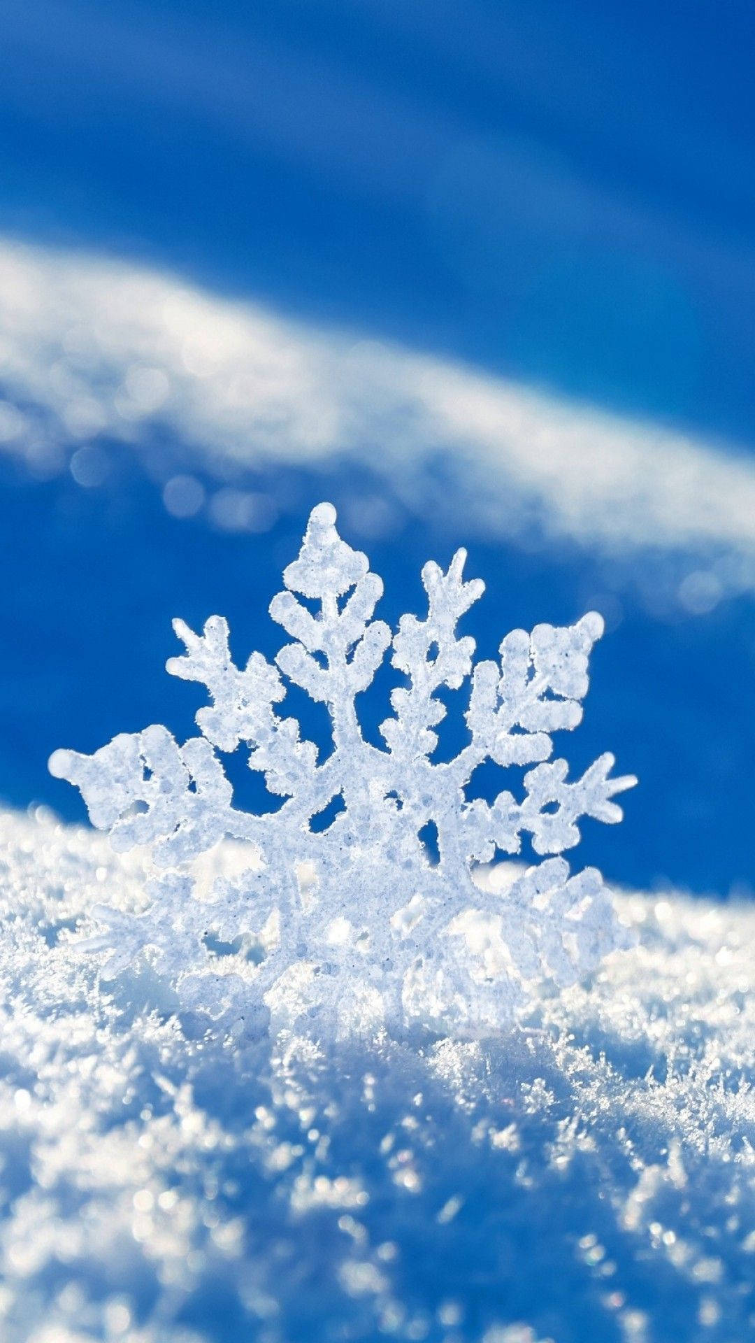 Snowflake iPhone 桌布 壁紙 — 一片精緻的雪花落在雪地上，背景是湛藍的天空和柔和的散景雲朵。