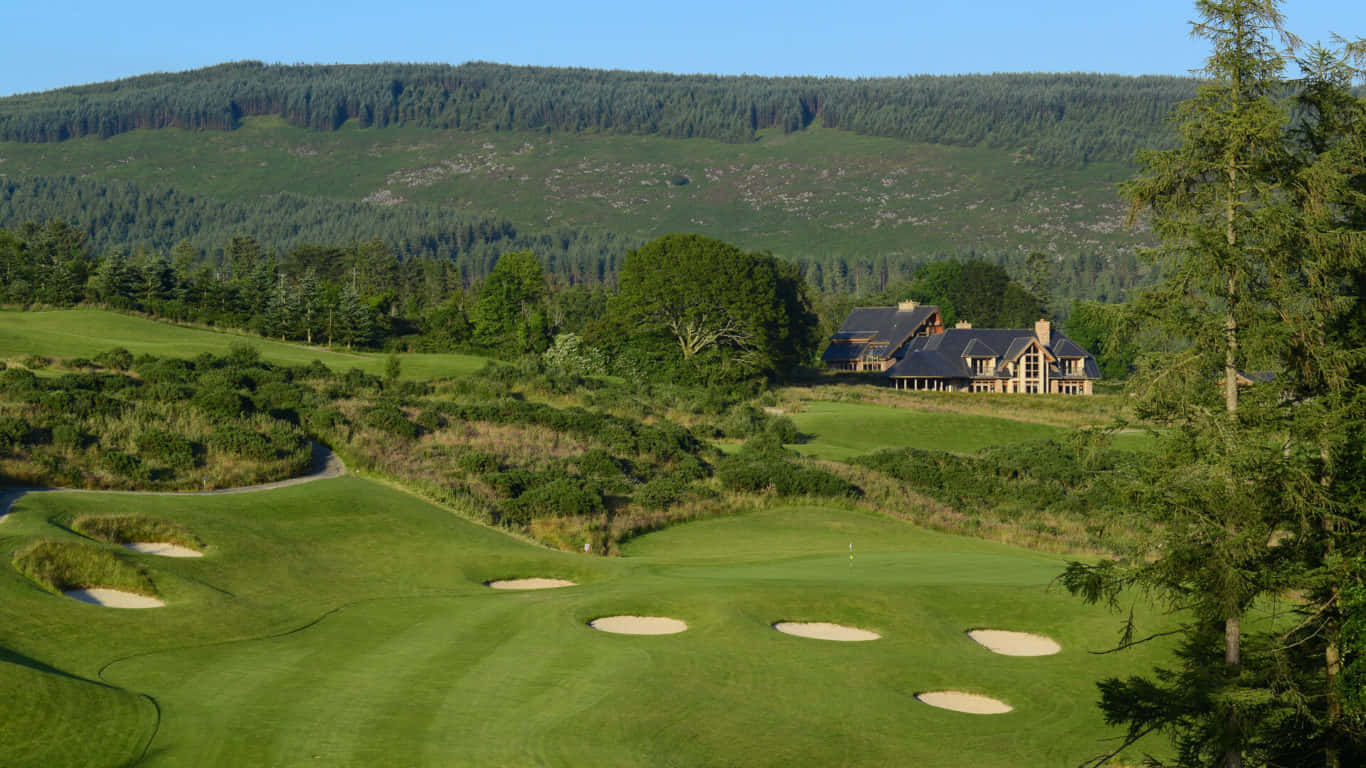 1366x768 Golf Course 桌布 壁紙 — 一片翠綠的高爾夫球場，設有沙坑，一座大房子隱藏在樹木之間，背景是森林覆蓋的山丘，天空清澈湛藍。