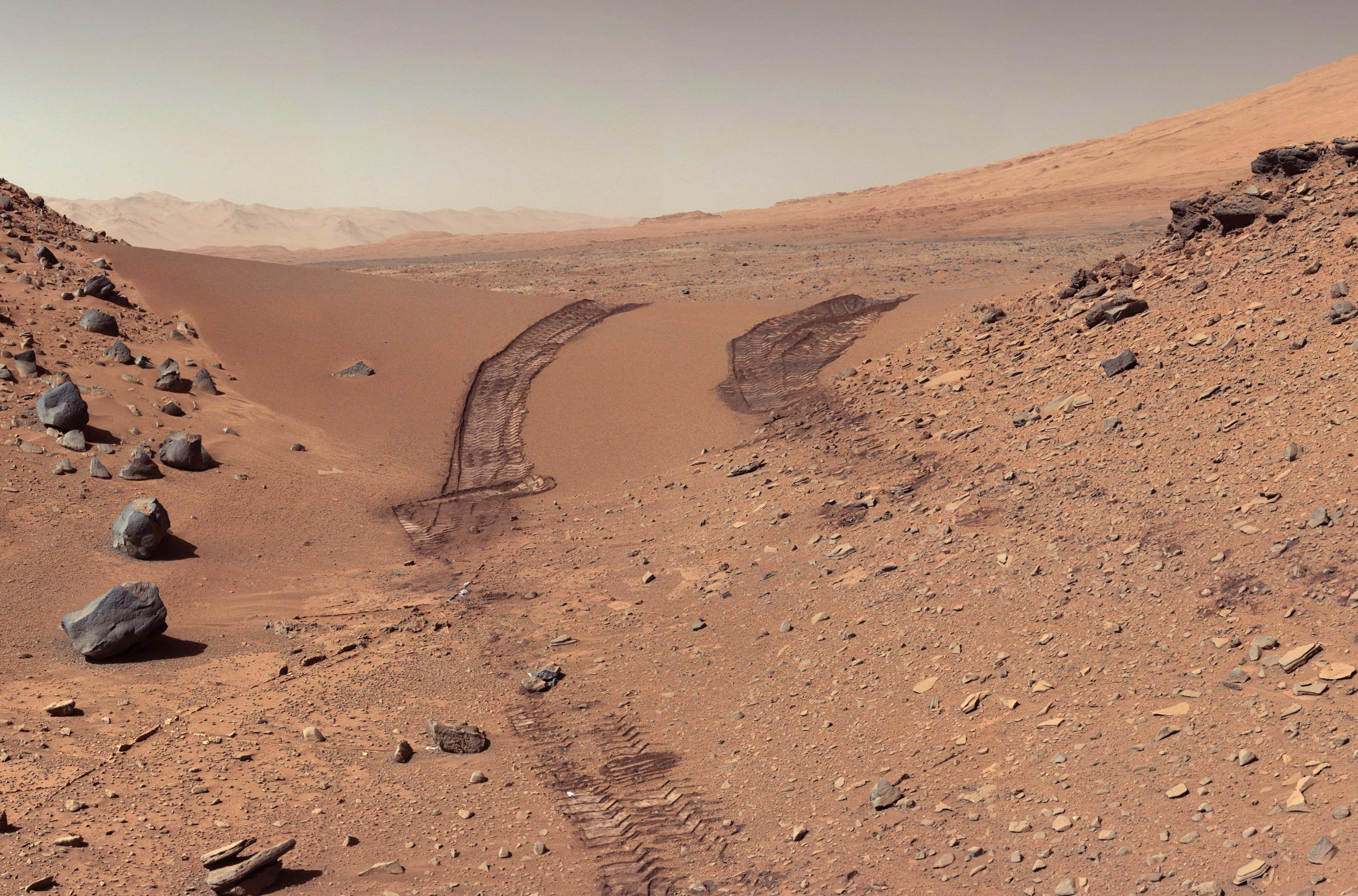 Mars Surface 桌布 壁紙 — 一片荒涼的紅色火星景觀，蜿蜒的探測車軌跡穿過塵土飛揚的沙丘和散落的岩石，天空朦朧。