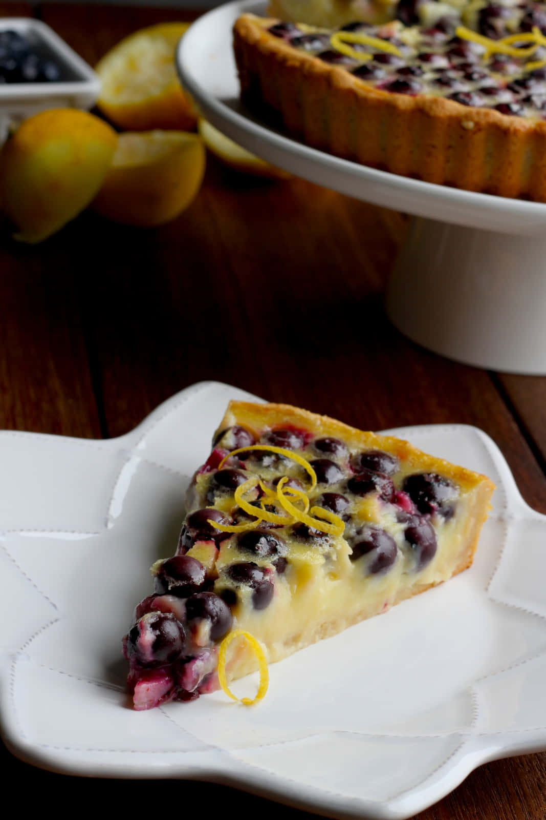 Blueberries Tart 桌布 壁紙 — 一片藍莓檸檬塔，飾以檸檬皮，放在白色盤子上，旁邊有整個檸檬和一個放在架上的完整塔。