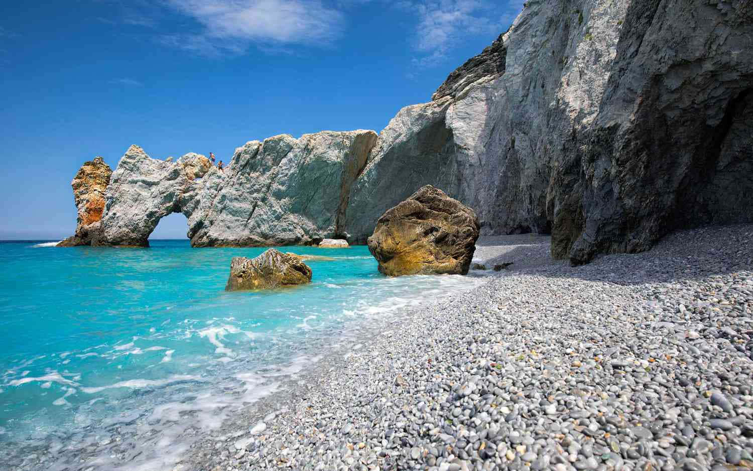 Greek Beach 桌布 壁紙 — 一片迷人的碧綠海水拍打著卵石海灘，在明亮的藍天下，由巨大的天然岩石拱門和高聳的懸崖構成框架。