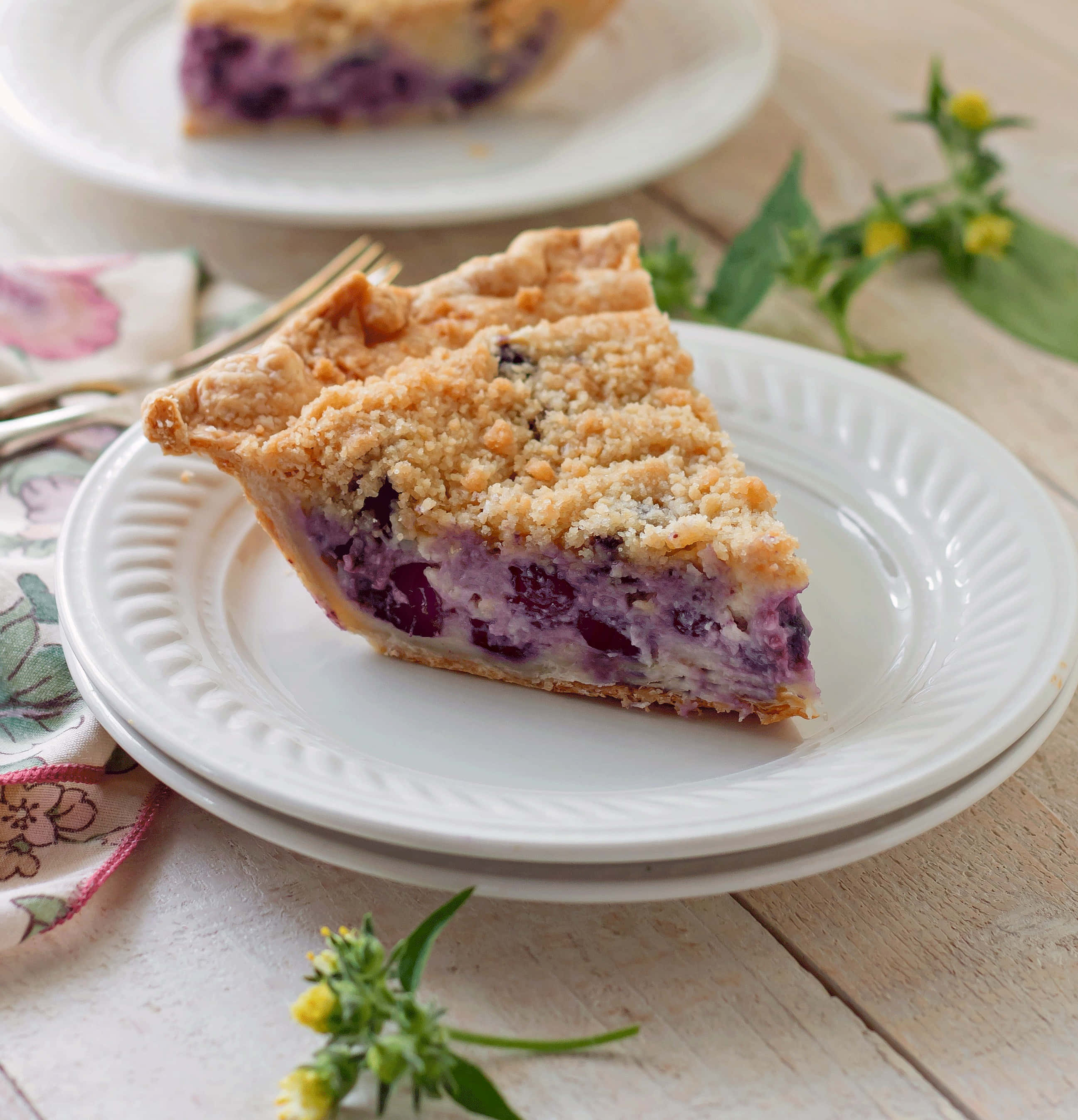 Blueberry Pie 桌布 壁紙 — 一片金黃脆皮的藍莓金寶派放在白色盤子上，置於鄉村風木桌上，旁邊有新鮮香草和花卉圖案餐巾。