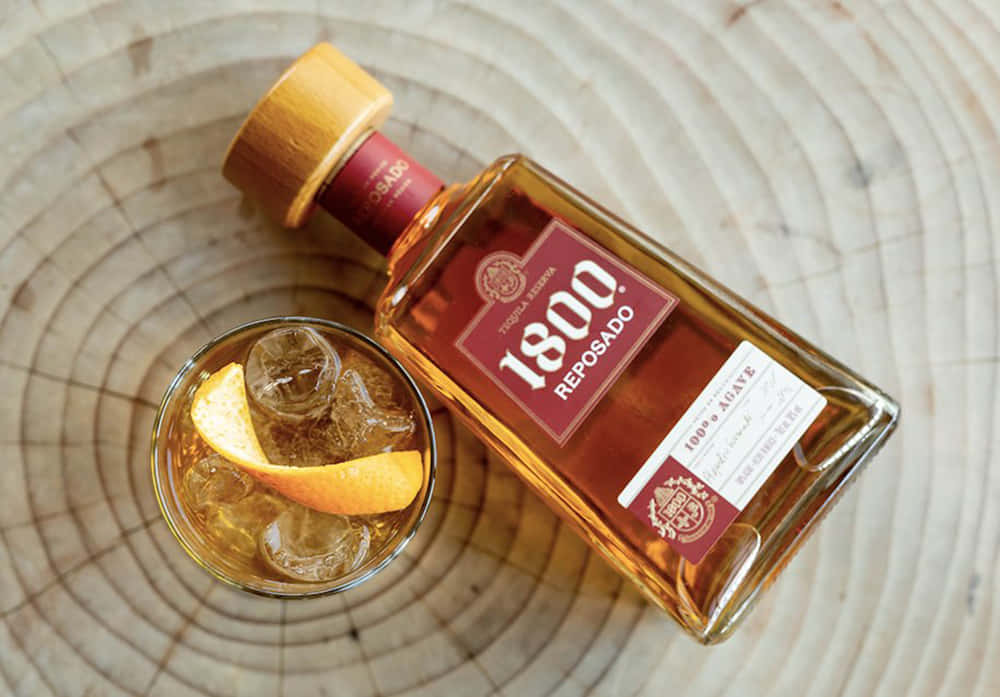 alcohol 桌布 壁紙 — 一瓶 1800 Reposado 龍舌蘭酒和一杯加冰和橙裝飾的玻璃杯放在一塊木切片上。
