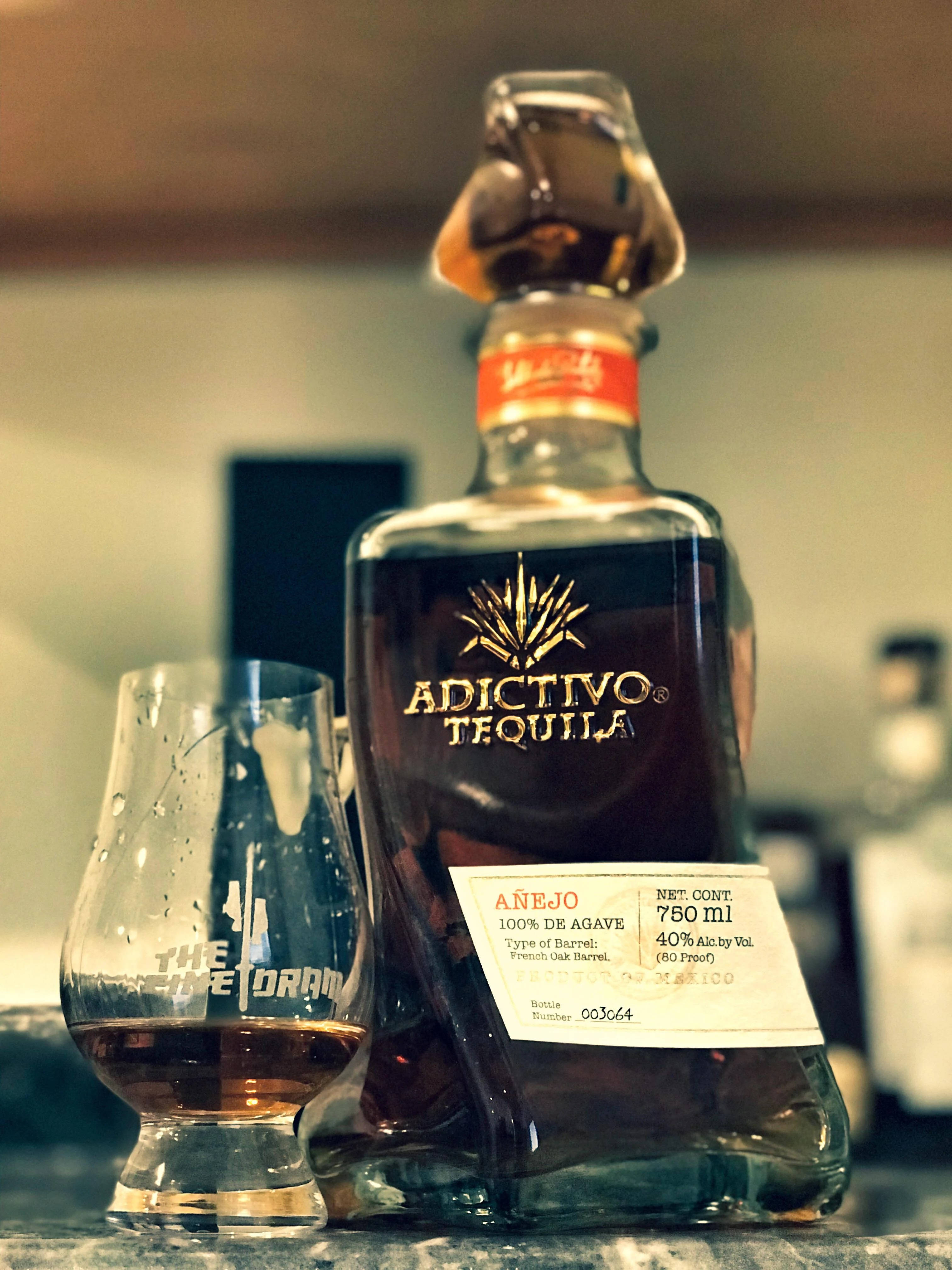 雙螢幕桌布 桌布 壁紙 — 一瓶 Adictivo Añejo 龍舌蘭酒放在吧檯上，旁邊有一個品酒杯，環境燈光溫暖。