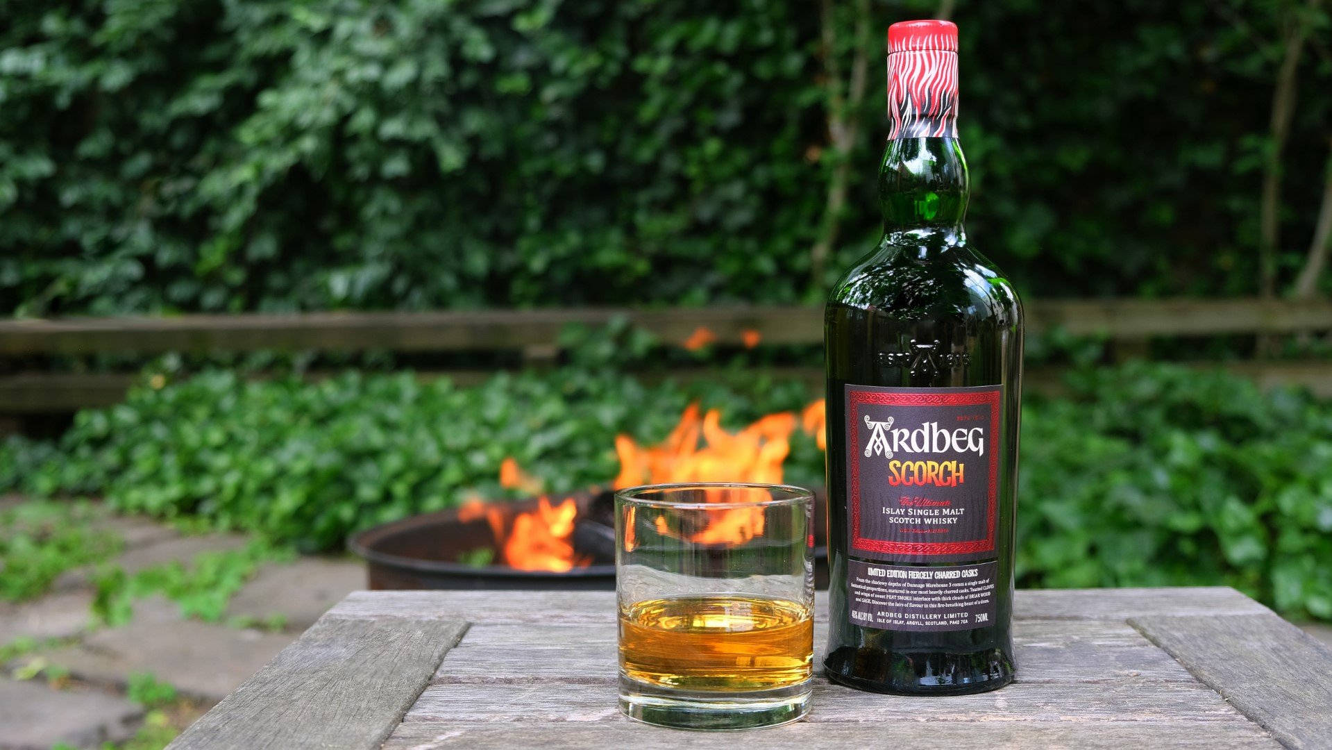 drinks 桌布 壁紙 — 一瓶 Ardbeg Scorch 威士忌和一個玻璃杯放在木桌上，旁邊是一個在茂密綠色花園中發光的火坑。