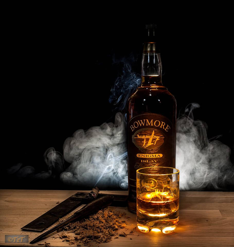 Whiskey 桌布 壁紙 — 一瓶 Bowmore Enigma 威士忌和酒杯在木桌上溫暖地發光，周圍有煙霧和雪茄剪。