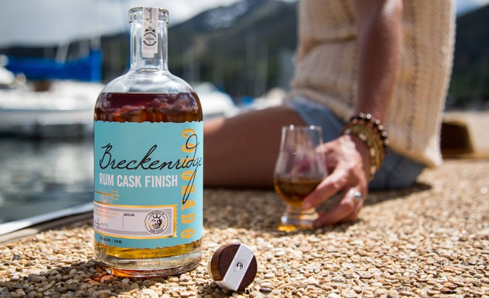 glass 桌布 壁紙 — 一瓶 Breckenridge Rum Cask Finish 威士忌放在鵝卵石上，旁邊有一個人拿著玻璃杯，背景是水、船隻和山脈。