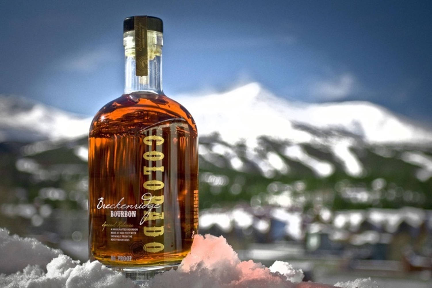 bottle 桌布 壁紙 — 一瓶 Breckenridge Colorado 波本威士忌放在新雪中，背景是白雪覆蓋的山脈和清澈的藍天。