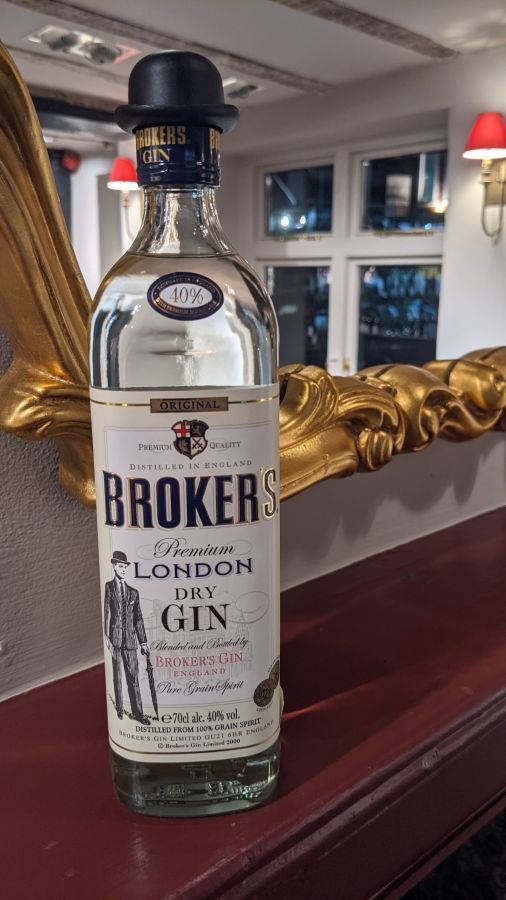 bottle 桌布 壁紙 — 一瓶 Brokers Gin 戴著高帽，放在紅色木質表面上，旁邊有一個華麗的金色鏡子，房間光線溫暖。