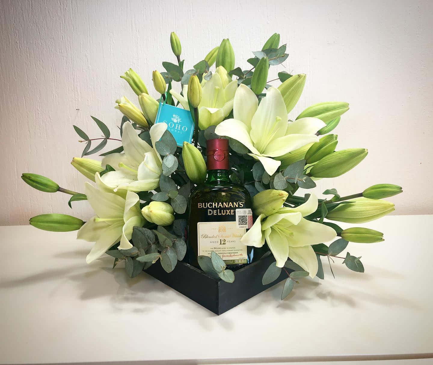 alcohol 桌布 壁紙 — 一瓶 Buchanan's Deluxe 威士忌，安放在一個黑色盒子裡，周圍是茂盛的白百合和綠色桉樹，置於白色表面上。