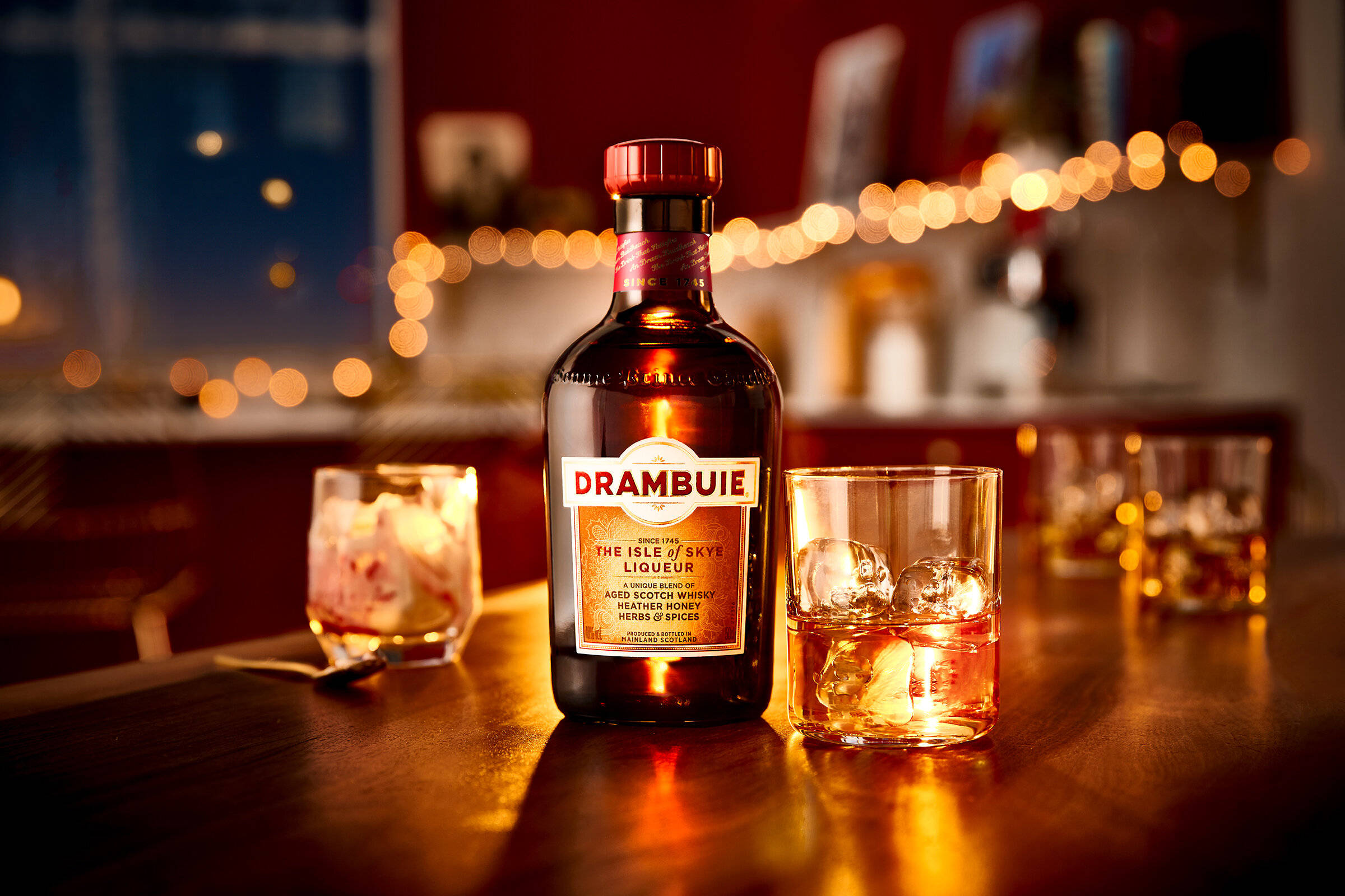 alcohol 桌布 壁紙 — 一瓶 Drambuie 利口酒和兩杯加冰的酒杯放在木製吧檯上，夜晚溫暖的串燈下。