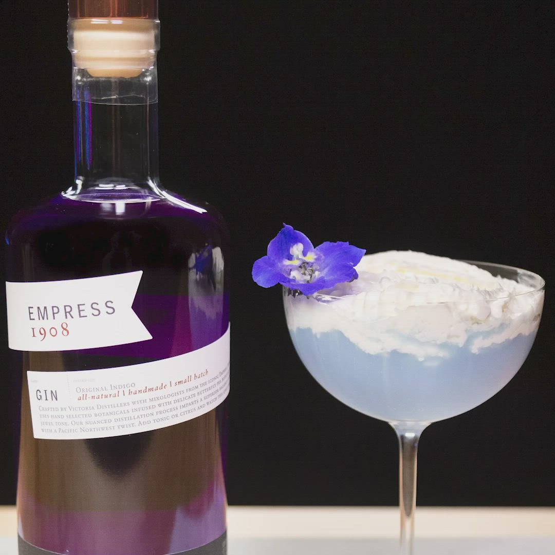cocktail 桌布 壁紙 — 一瓶 Empress 1908 紫色琴酒放在一杯淺藍色雞尾酒旁邊，雞尾酒上配有鮮奶油和一朵紫色花，背景為黑色。