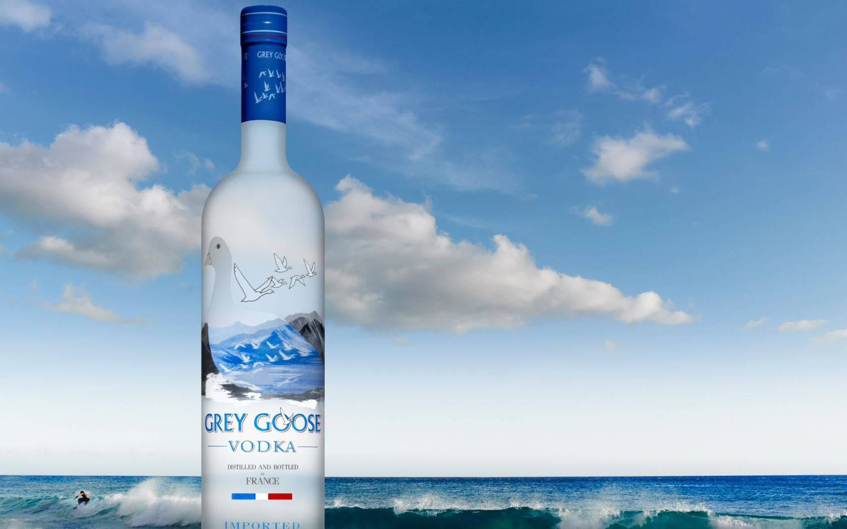 飲料 桌布 壁紙 — 一瓶 Grey Goose 伏特加酒在明亮的藍天、蓬鬆的雲朵和下方翻騰的海浪襯托下高高聳立。