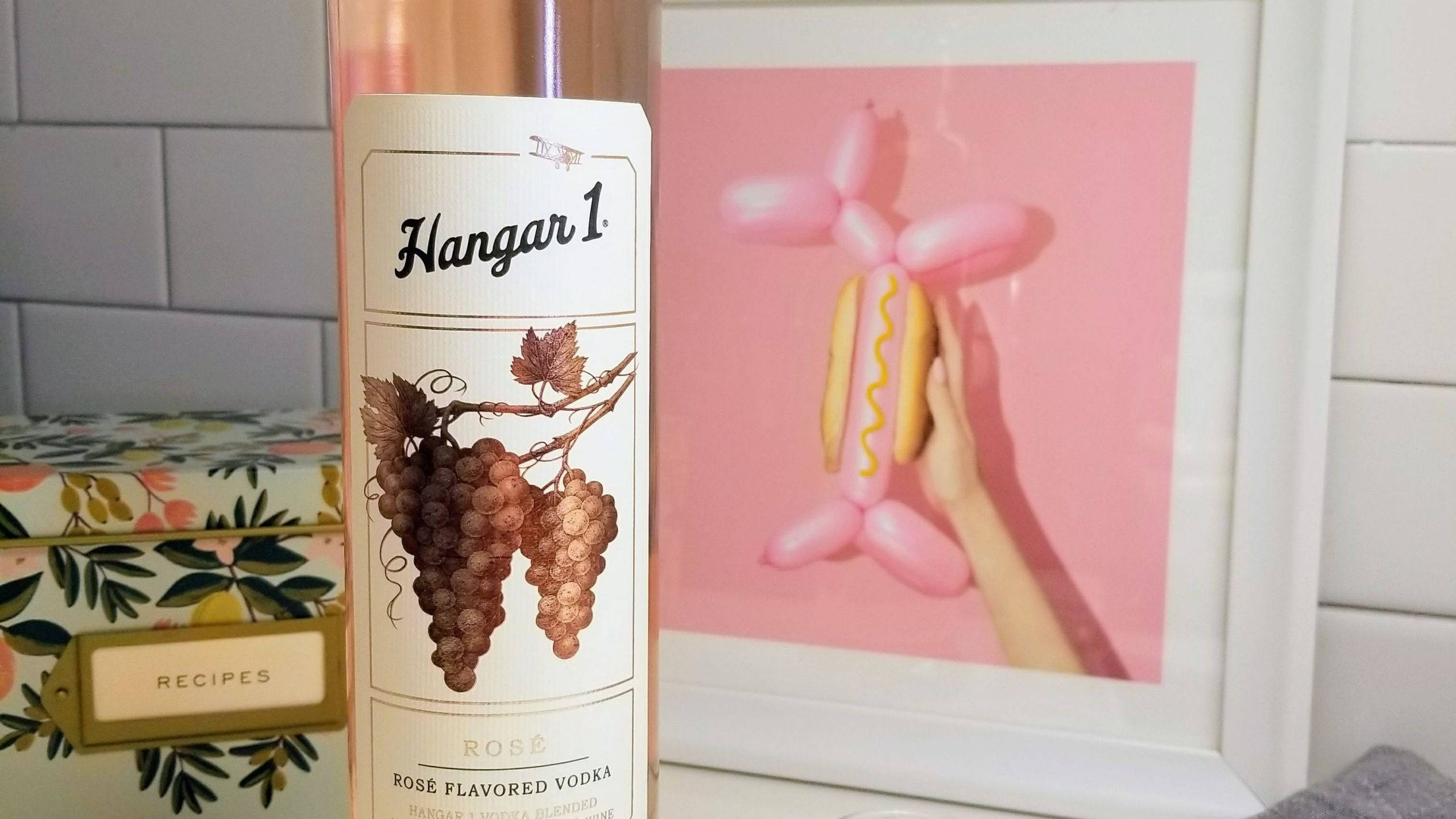 bottle 桌布 壁紙 — 一瓶 Hangar 1 Rose Flavored Vodka 放在一個相框旁邊，相框裡是一隻手拿著粉紅色氣球狗，背景為粉紅色。