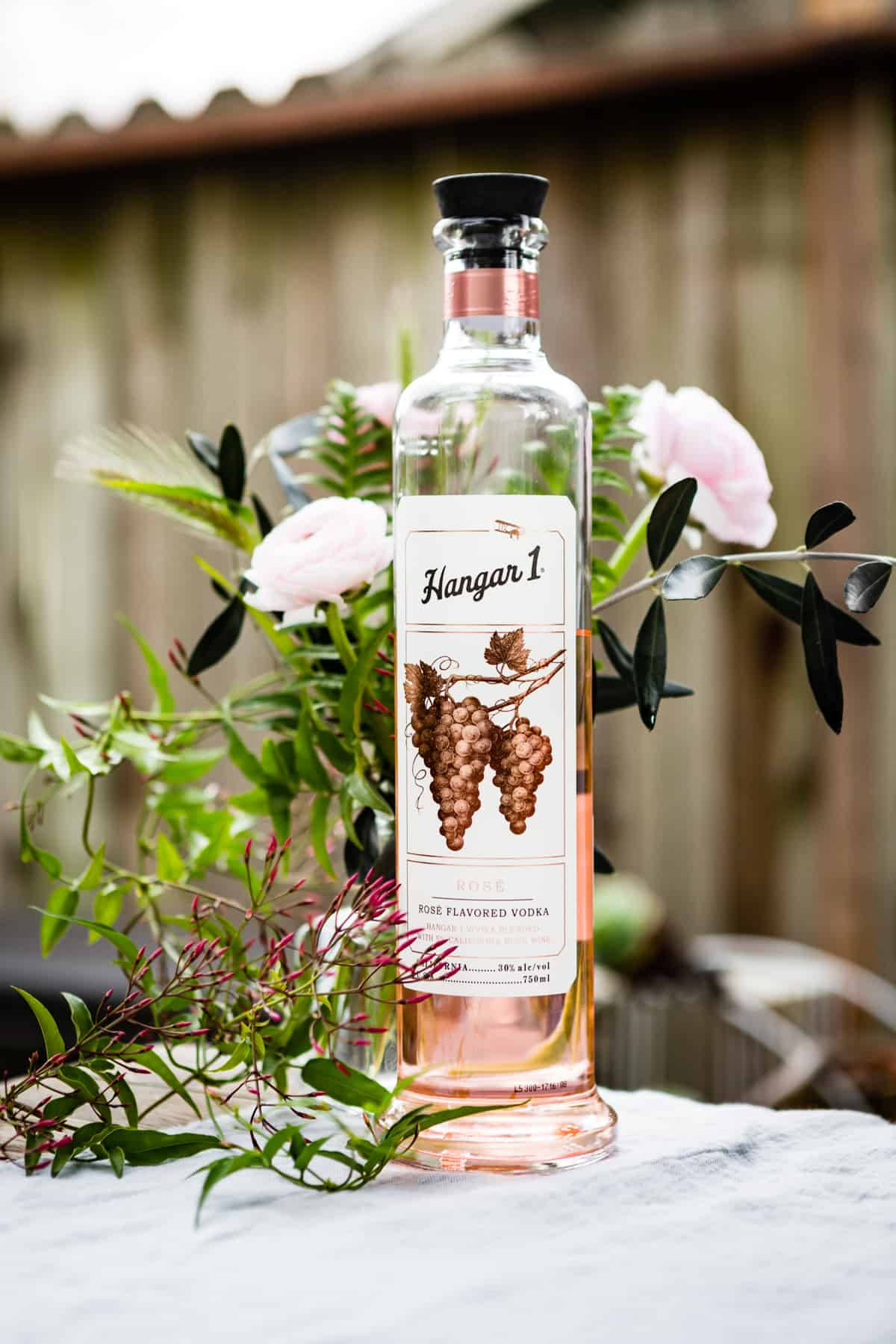bottle 桌布 壁紙 — 一瓶 Hangar 1 Rose Flavored Vodka 優雅地放在大理石表面上，周圍環繞著柔和的粉紅色花朵和綠色植物。