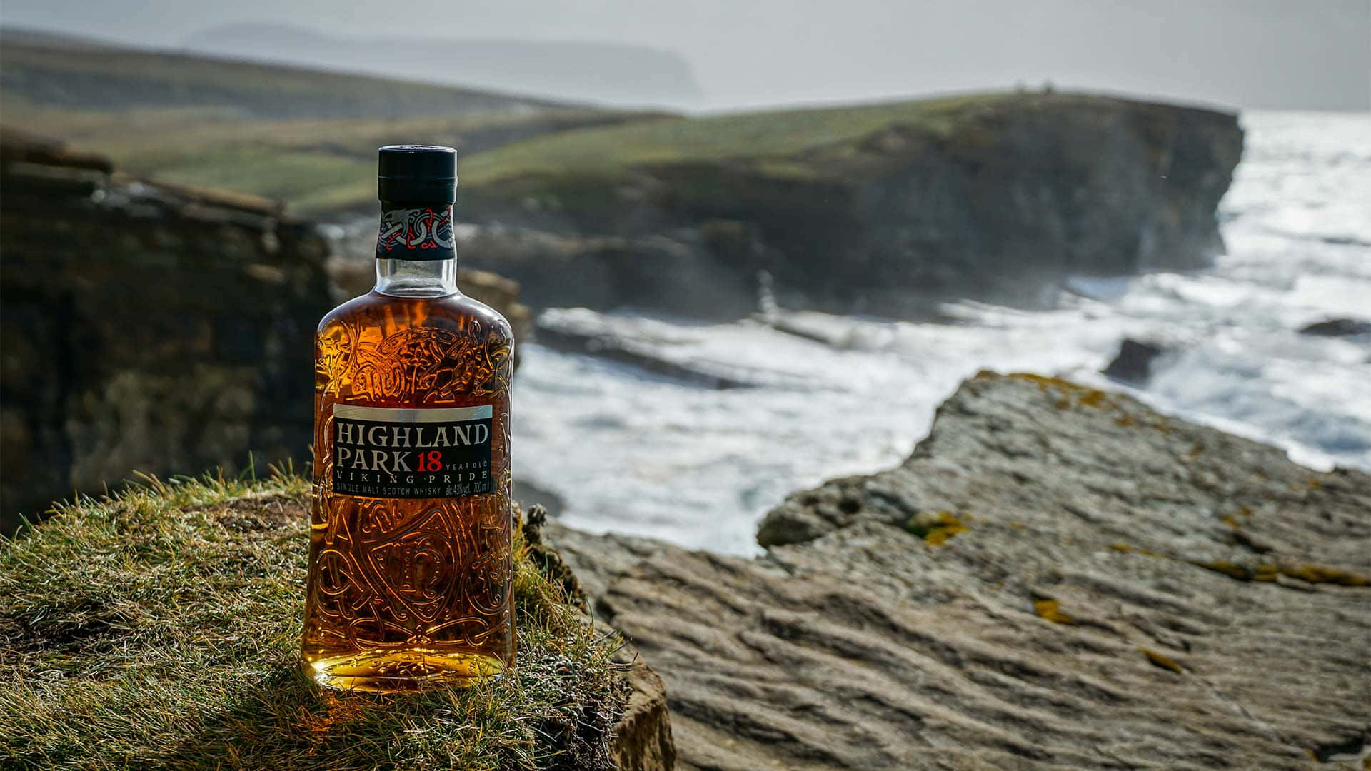 alcohol 桌布 壁紙 — 一瓶 Highland Park 18 年威士忌矗立在長滿苔蘚的懸崖上，俯瞰著洶湧的海浪和霧氣瀰漫的海岸懸崖。