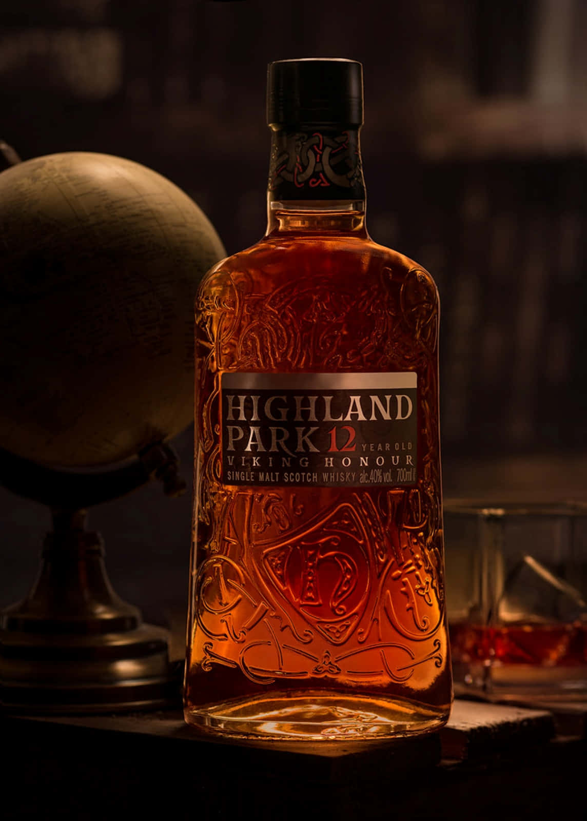 bottle 桌布 壁紙 — 一瓶 Highland Park 12 Year Old Viking Honour 威士忌在昏暗的燈光下，在地球儀和玻璃杯旁邊溫暖地發光。