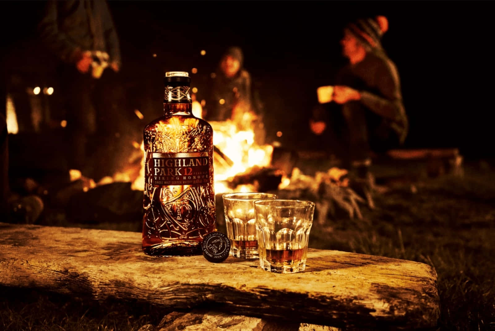 booze 桌布 壁紙 — 一瓶 Highland Park 12 威士忌和兩個酒杯在夜間噼啪作響的營火旁的木頭上溫暖地發光。