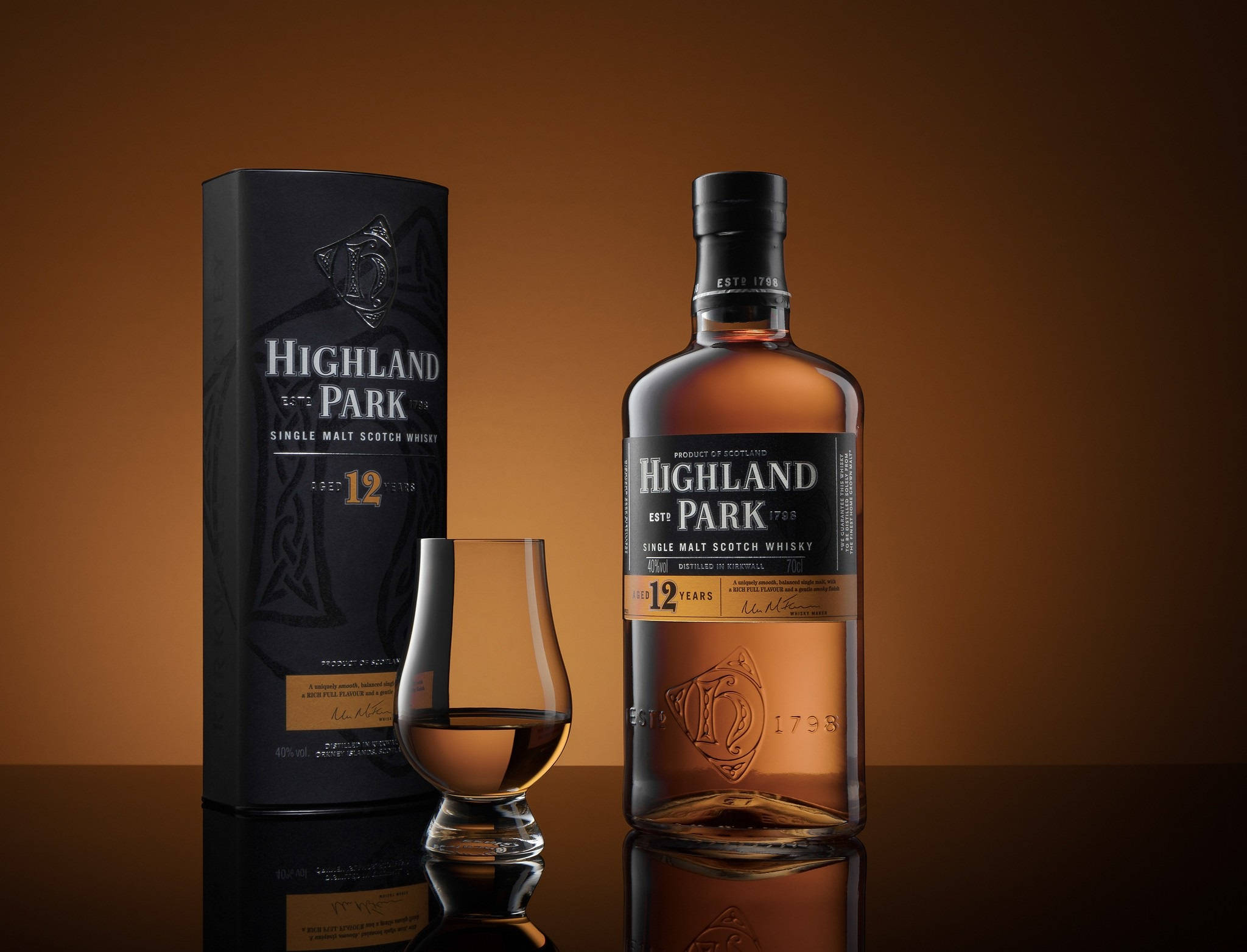 booze 桌布 壁紙 — 一瓶 Highland Park 12 年單一麥芽蘇格蘭威士忌，旁邊放著它的盒子和一個品酒杯，放在反射表面上，背景是溫暖的棕色漸變。