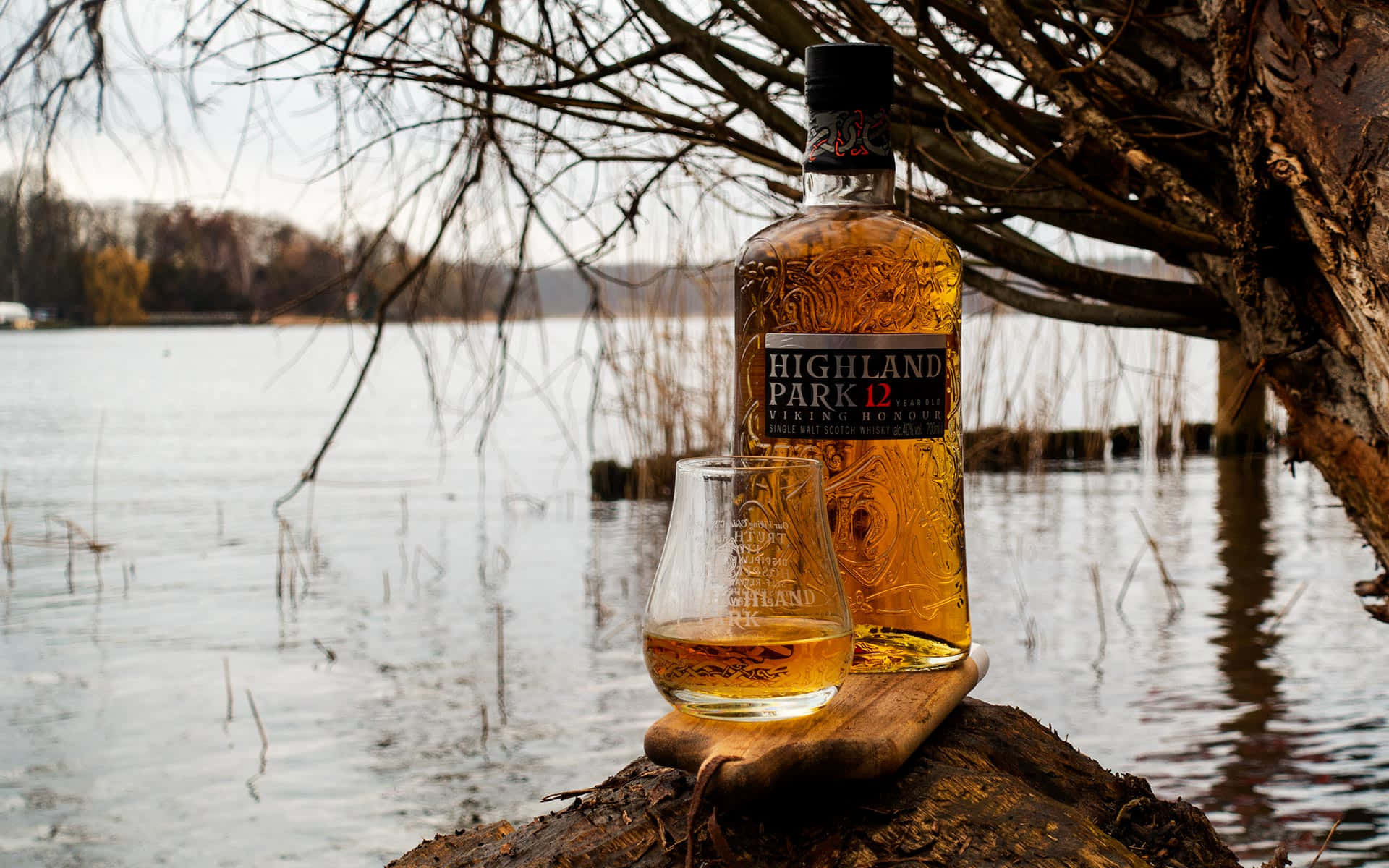 booze 桌布 壁紙 — 一瓶 Highland Park 12 Viking Honour 威士忌和酒杯放在平靜湖邊的木頭上，被光禿的冬季樹枝環繞。