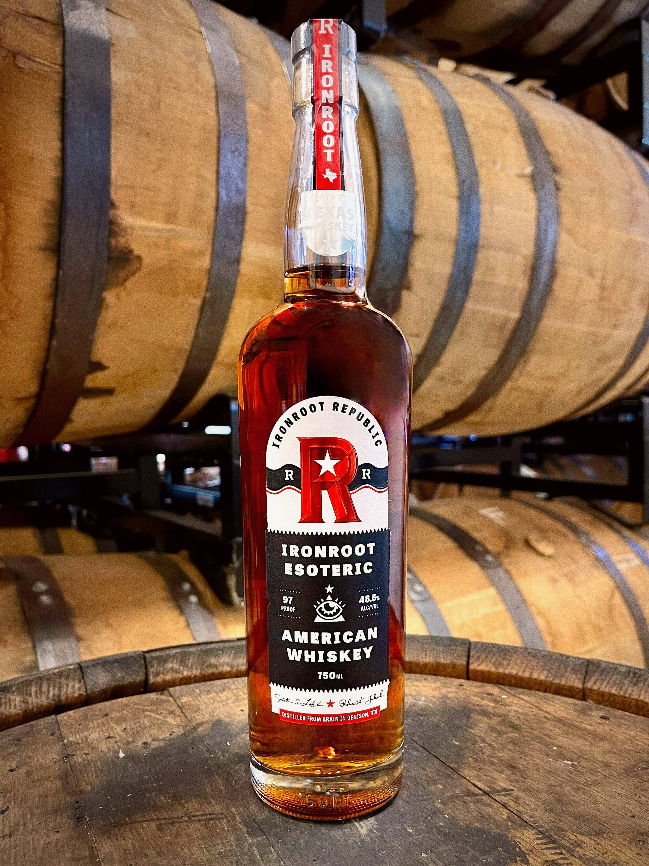 飲料 桌布 壁紙 — 一瓶 Ironroot Esoteric American Whiskey 放在一個木桶上，背景是鄉村風格的釀酒廠，排列著正在陳年的威士忌酒桶。