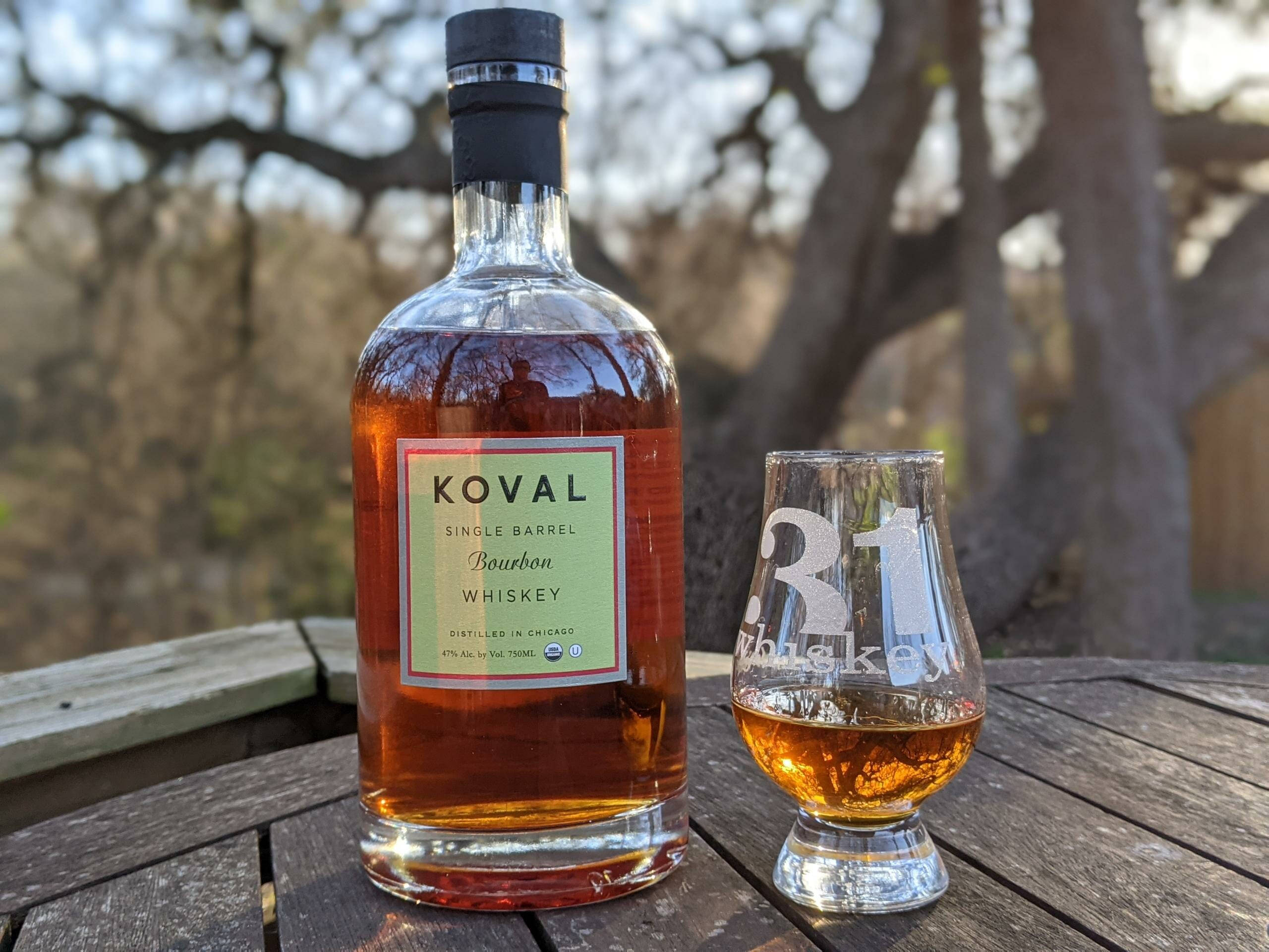 alcohol 桌布 壁紙 — 一瓶 Koval 單桶波本威士忌和一個盛滿琥珀色液體的品牌酒杯，放在戶外一張風化的木桌上，周圍是光禿禿的樹木，在柔和的光線下。
