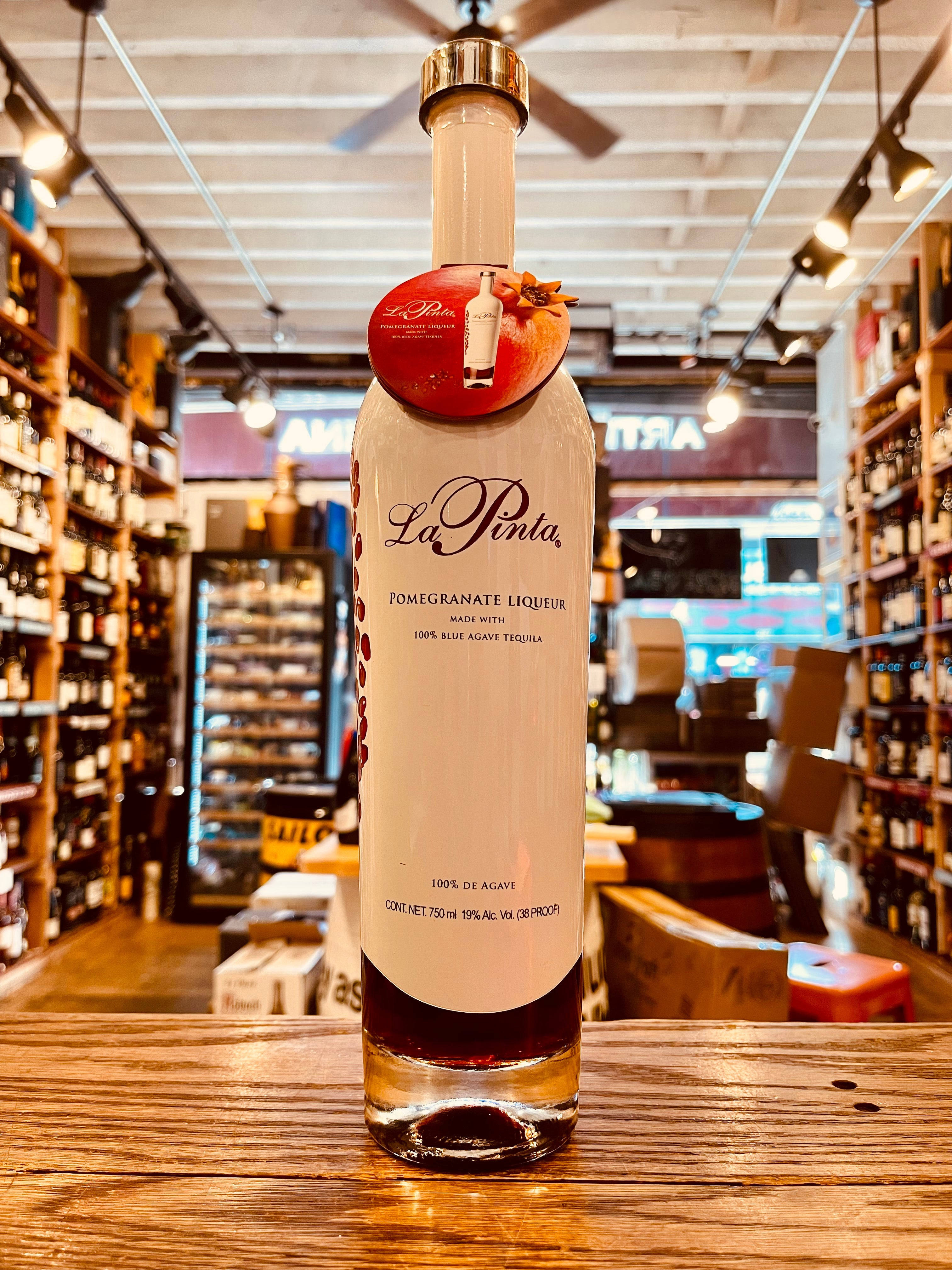 bottle 桌布 壁紙 — 一瓶 La Pinta 石榴利口酒放在一間燈光溫暖的酒鋪木檯中央，背景是擺滿酒瓶的貨架。