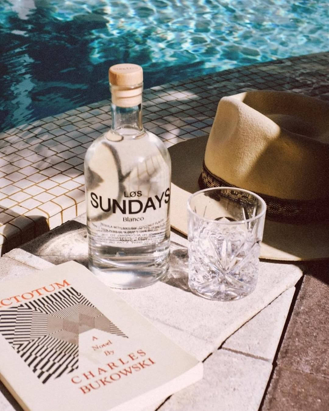 alcohol 桌布 壁紙 — 一瓶 Los Sundays Blanco 龍舌蘭酒、玻璃杯和軟呢帽放在陽光普照的泳池邊瓷磚上，旁邊有一本書。