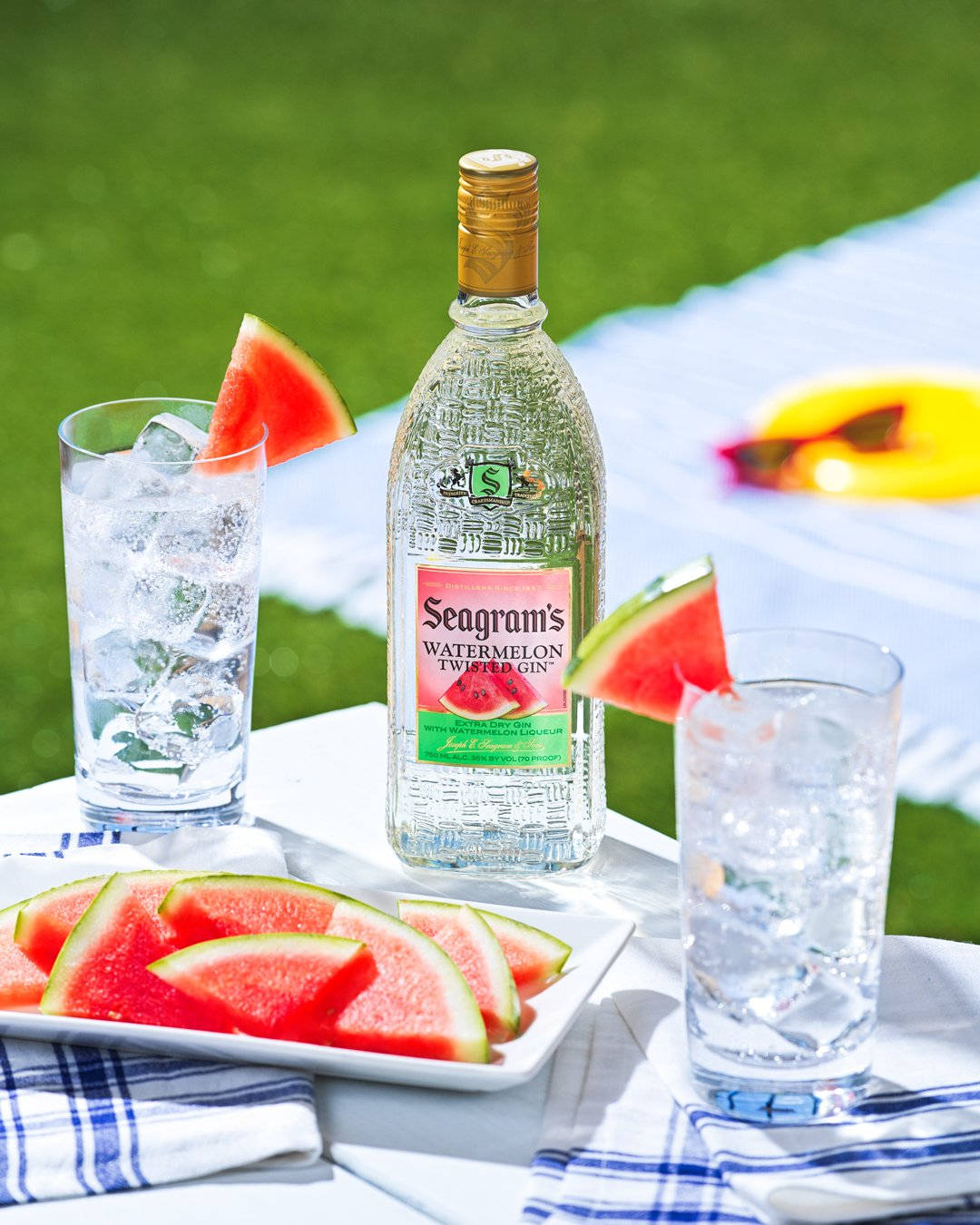 動態圖 桌布 壁紙 — 一瓶 Seagram's Watermelon Twisted Gin 放在白色桌布上，旁邊有兩個杯子和西瓜片，在陽光明媚的草地上。