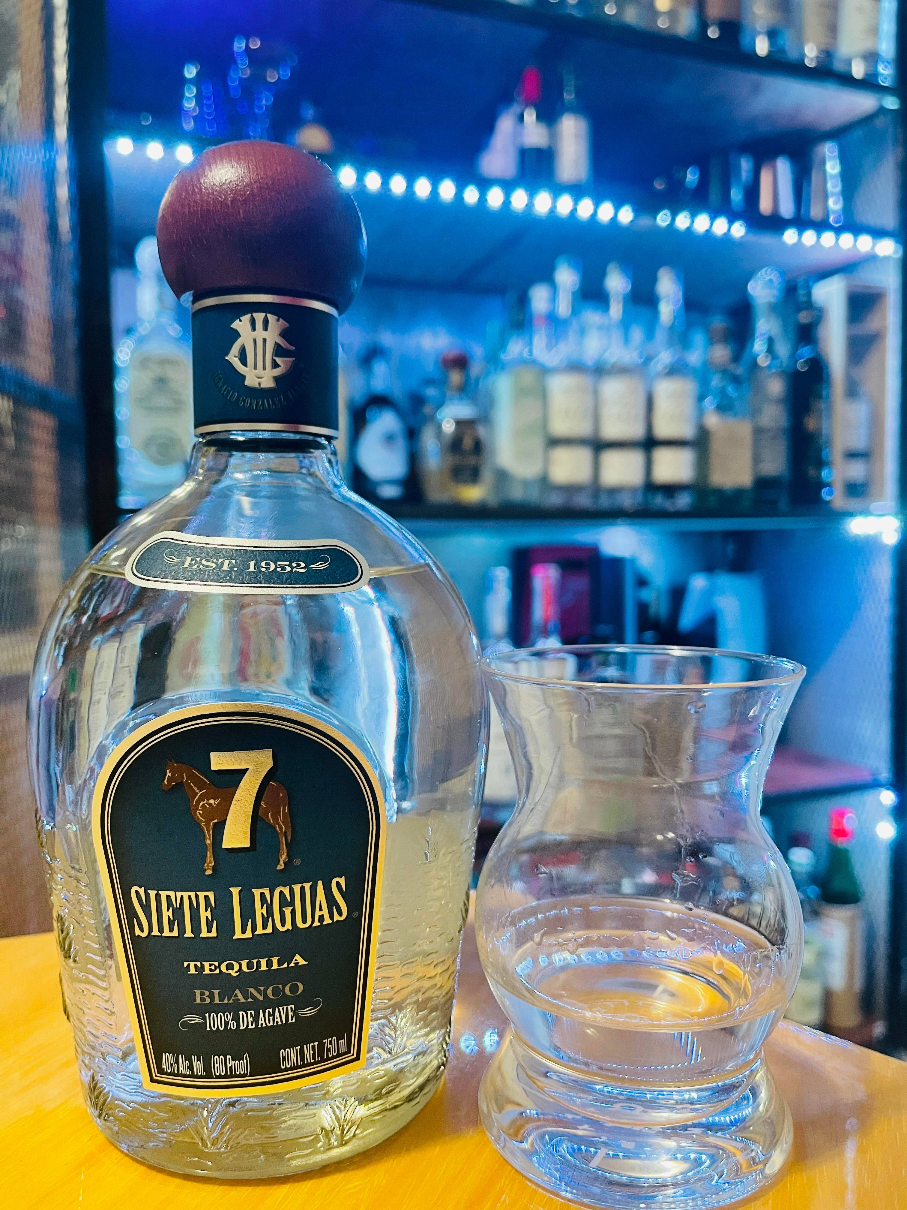 手機 桌布 壁紙 — 一瓶 Siete Leguas Blanco 龍舌蘭酒放在木製吧檯上，旁邊有一個玻璃杯，背景是藍色燈光照亮的酒架。