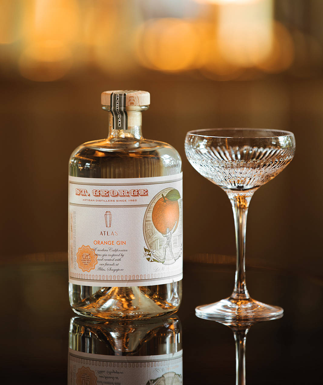 bottle 桌布 壁紙 — 一瓶 St George Atlas Orange Gin 放在深色反光表面上，旁邊有一個水晶 coupe 杯，背景有溫暖的散景燈光。