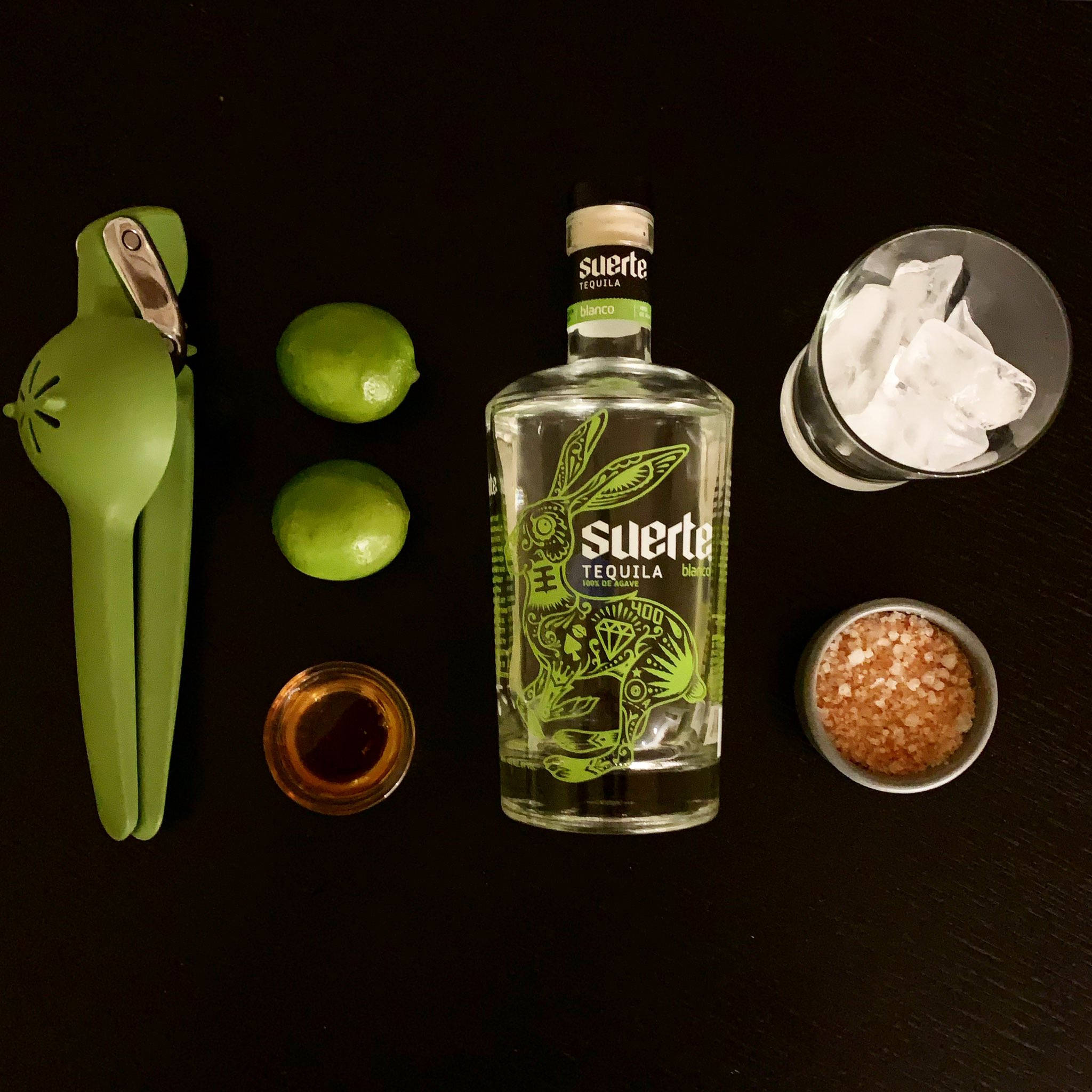 glass 桌布 壁紙 — 一瓶 Suerte 龍舌蘭酒，周圍環繞著萊姆、萊姆壓榨器、鹽和冰塊，置於深色木質表面上。