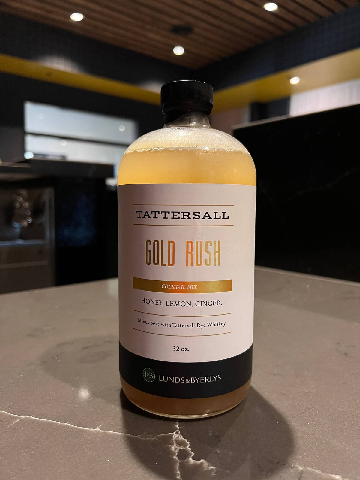 飲料 桌布 壁紙 — 一瓶 Tattersall Gold Rush 雞尾酒調配液放在現代酒吧的深色大理石檯面上，周圍有溫暖的環境燈光。