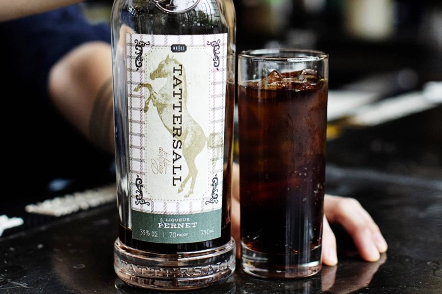 飲料 桌布 壁紙 — 一瓶 Tattersall Pernot 利口酒和一杯加冰的深色汽水放在深色吧檯上，旁邊有一隻手擱著。