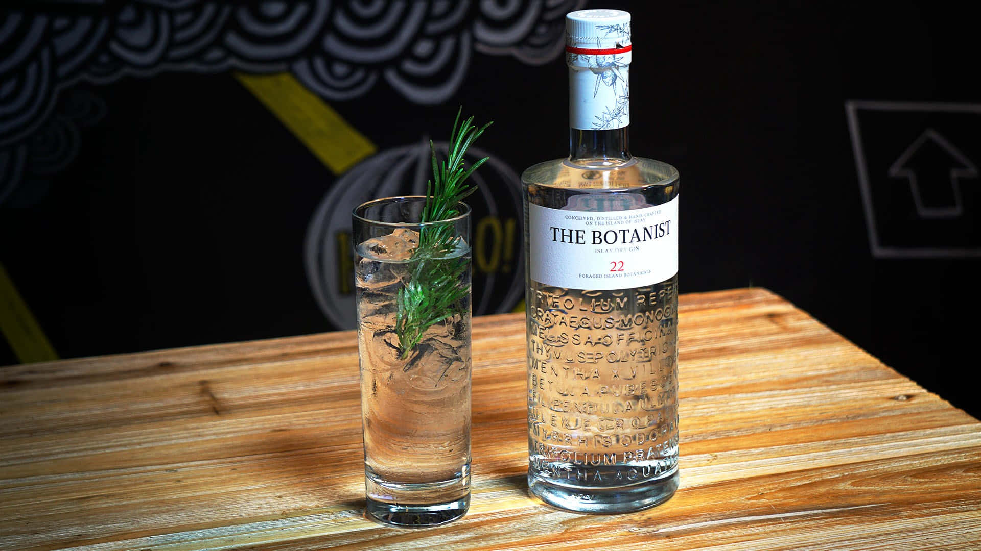 drinks 桌布 壁紙 — 一瓶 The Botanist 琴酒和一杯加冰和迷迭香的酒杯放在木桌上，背景是深色黑板牆。