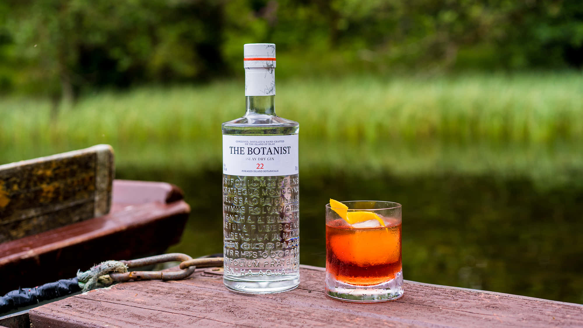 cocktail 桌布 壁紙 — 一瓶 The Botanist 琴酒和一杯帶有橙片的雞尾酒放在平靜綠色池塘邊的風化木碼頭上。