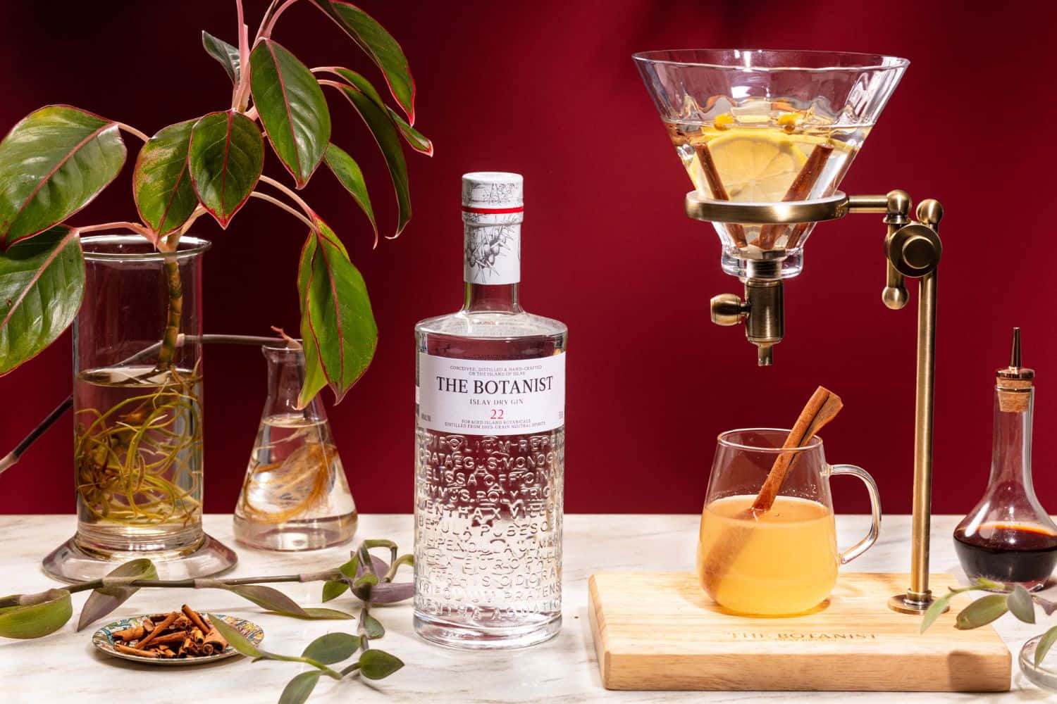 drinks 桌布 壁紙 — 一瓶 The Botanist 琴酒放在一杯植物雞尾酒旁邊，雞尾酒配有肉桂棒和檸檬片，周圍環繞著香草和實驗室玻璃器皿，置於大理石表面上，背景為深紅色。