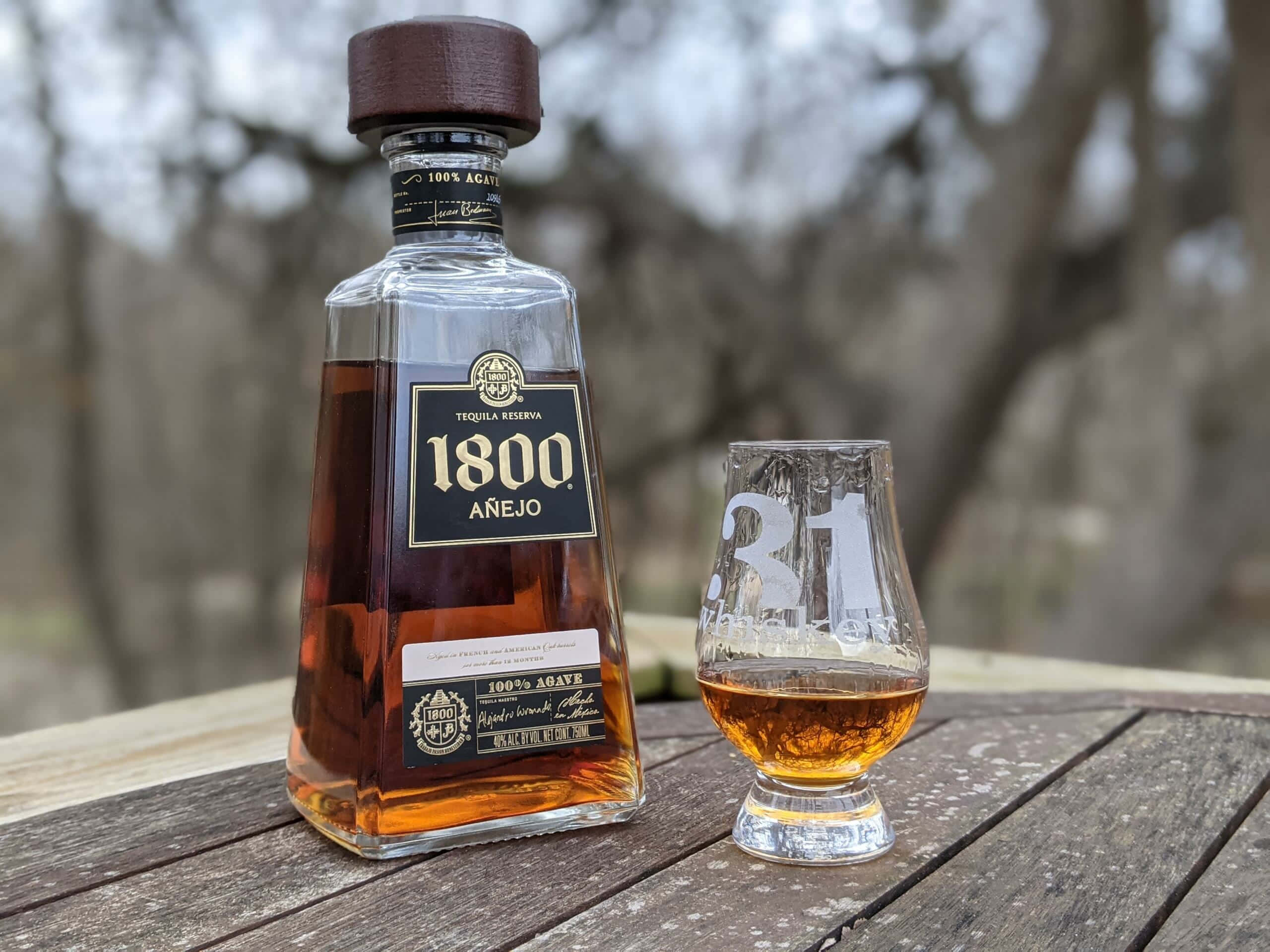 alcohol 桌布 壁紙 — 一瓶1800 Añejo龍舌蘭酒和一個品酒杯放在戶外一張風化的木桌上，背景是模糊的樹木。