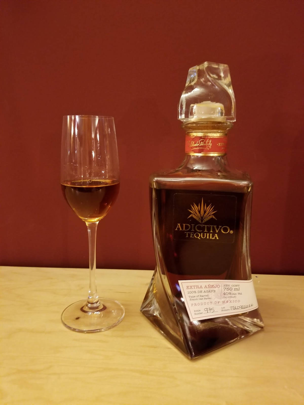 高清 桌布 壁紙 — 一瓶Adictivo Tequila Extra Añejo放在木質表面上，旁邊有一個裝滿酒的玻璃杯，背景是深紅色牆壁。