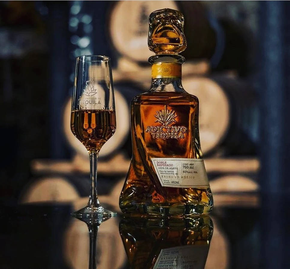 iPhone 桌布 壁紙 — 一瓶Adictivo Tequila和一個裝滿的玻璃杯在溫暖燈光下閃爍，背景是木桶。