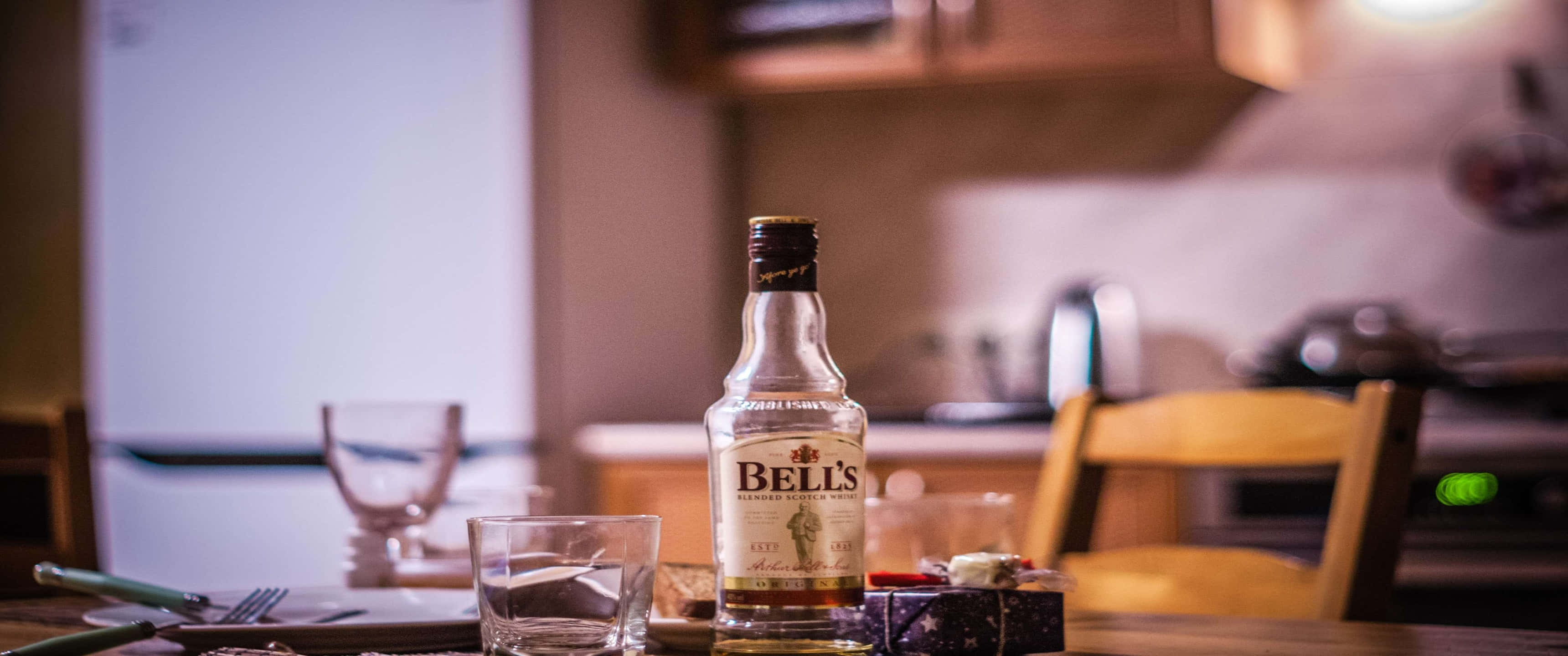 Kitchen 桌布 壁紙 — 一瓶Bell's威士忌放在一張木桌上，廚房燈光溫暖，背景元素模糊。