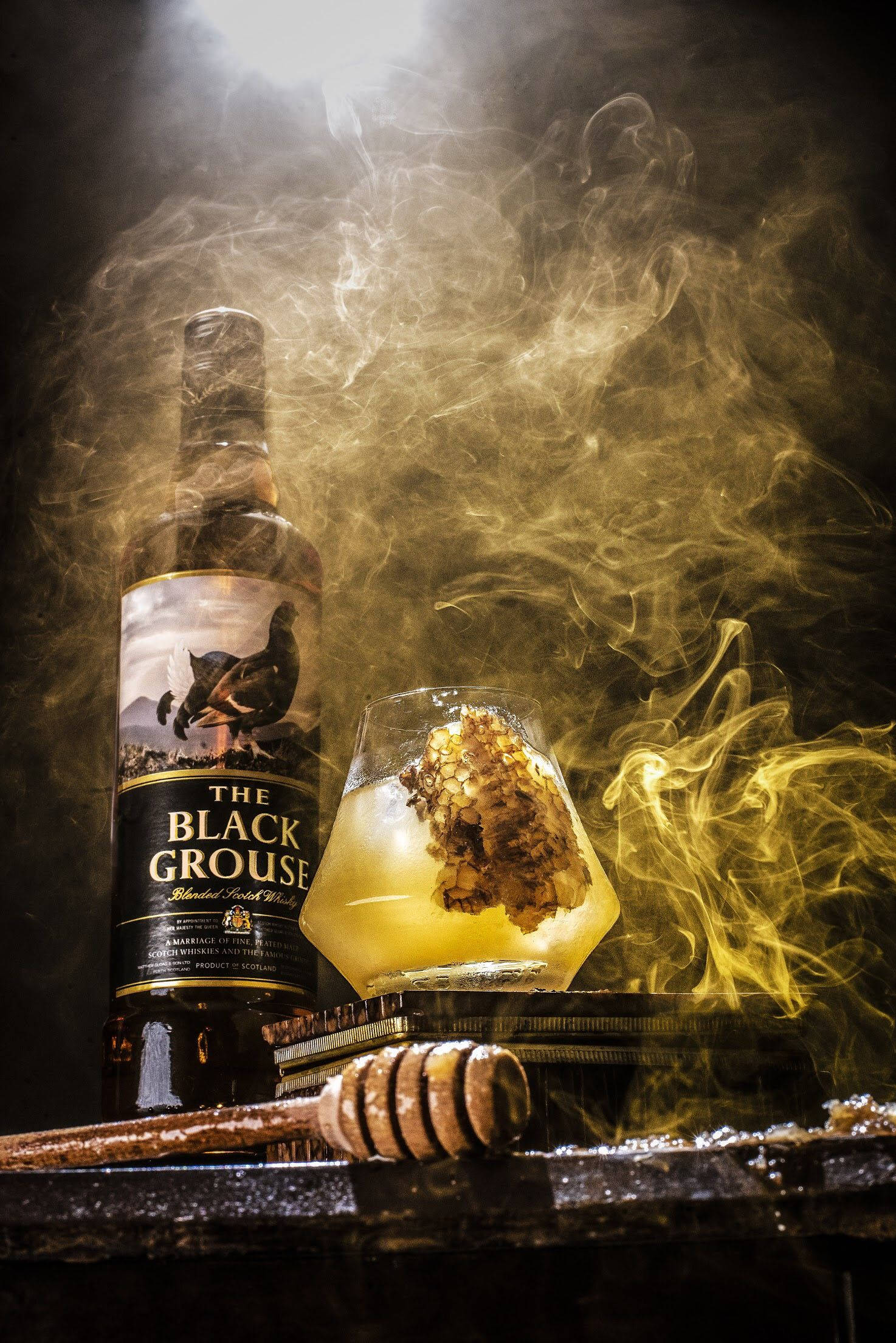 Whiskey 桌布 壁紙 — 一瓶Black Grouse威士忌和一個裝滿蜂巢的玻璃杯在質樸的木質表面上，於旋轉的金色煙霧中溫暖地閃耀。
