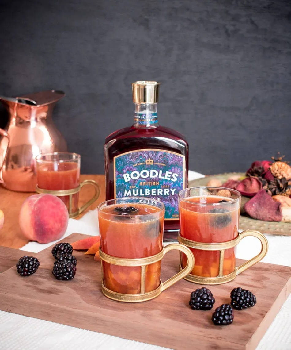 Peach 桌布 壁紙 — 一瓶Boodles Mulberry Gin旁邊有兩個金色杯子，裝滿了莓果浸泡的飲料，放在木板上。