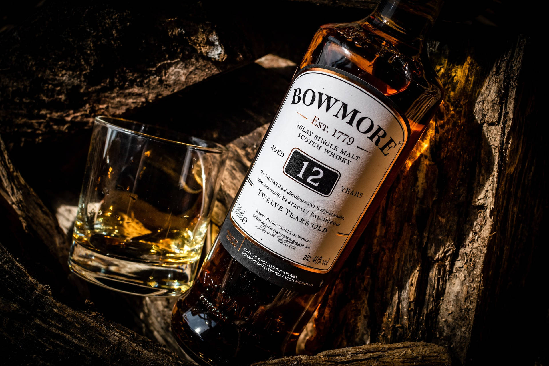 Whiskey 桌布 壁紙 — 一瓶Bowmore 12年威士忌和酒杯放在一個深色木桶中，帶有溫暖的金色燈光。
