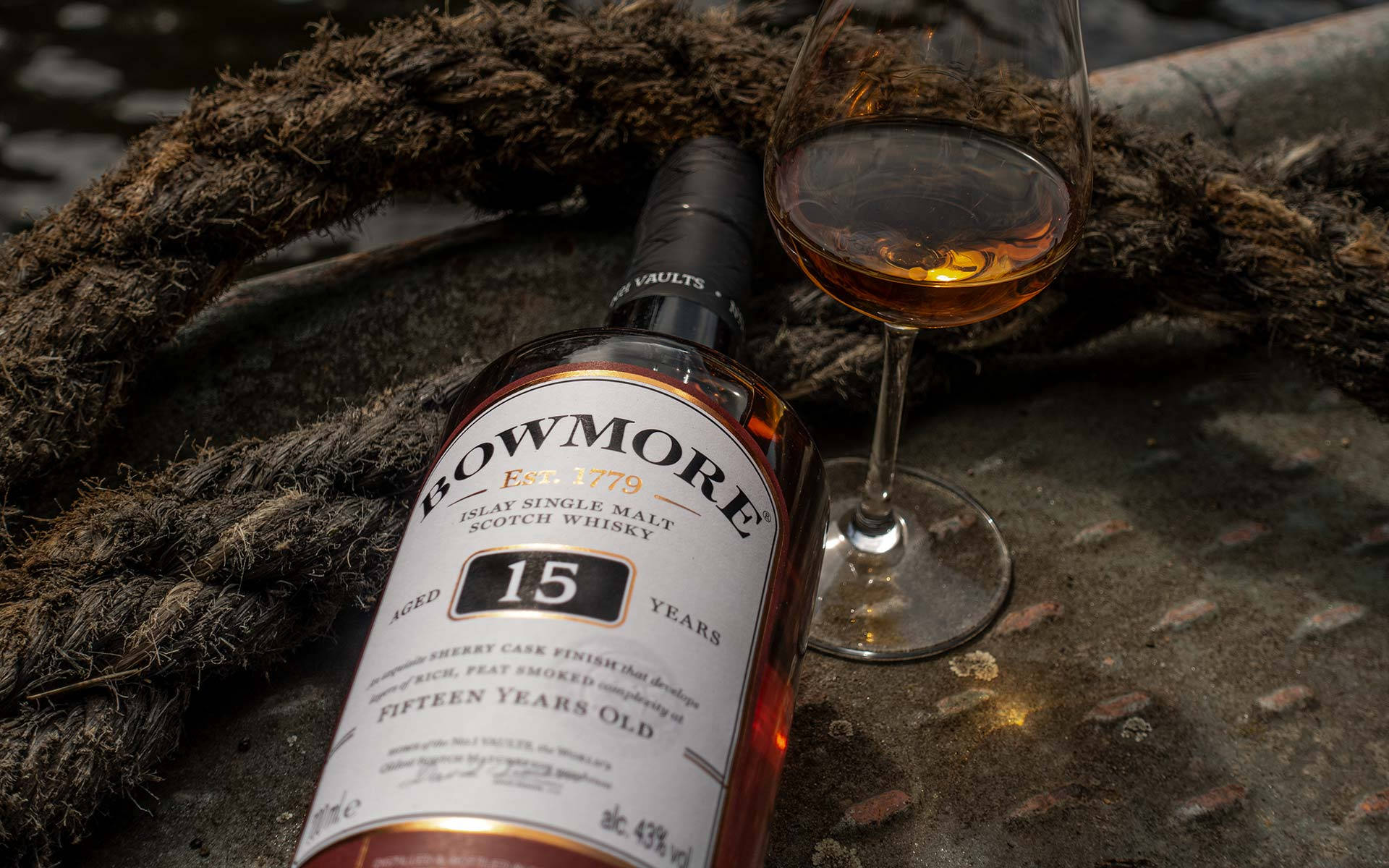 Whiskey 桌布 壁紙 — 一瓶Bowmore 15年艾雷島單一麥芽威士忌放在一個裝滿酒的玻璃杯旁邊，置於被粗繩纏繞的風化木質表面上。