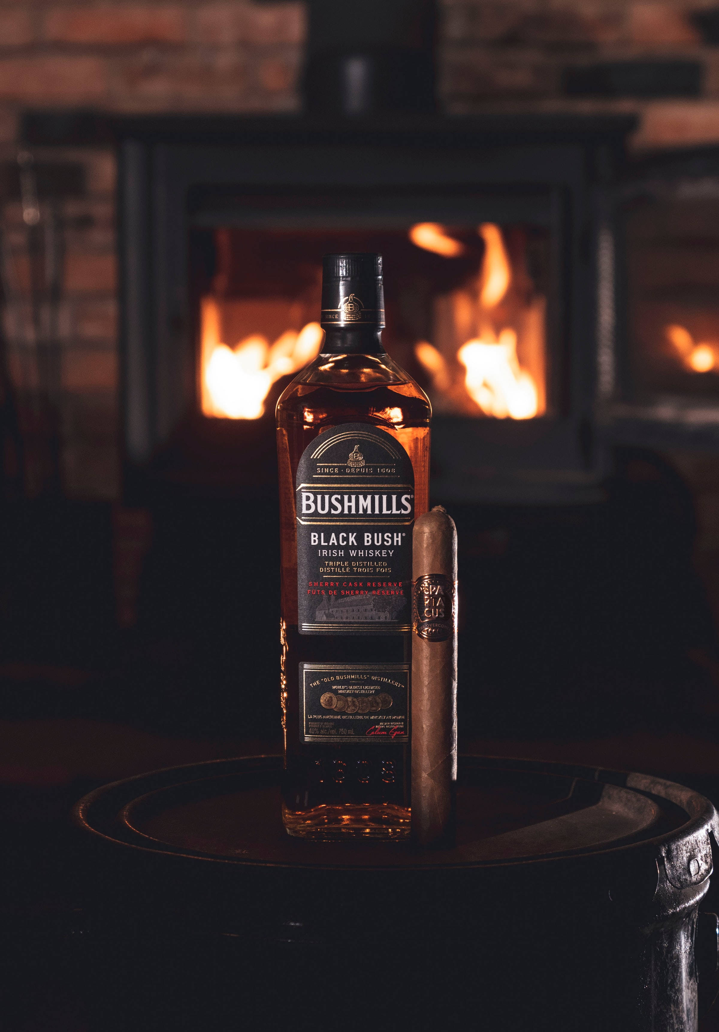 alcohol 桌布 壁紙 — 一瓶Bushmills Black Bush愛爾蘭威士忌和一支雪茄放在一個木桶上，前面是昏暗房間裡發光的壁爐。