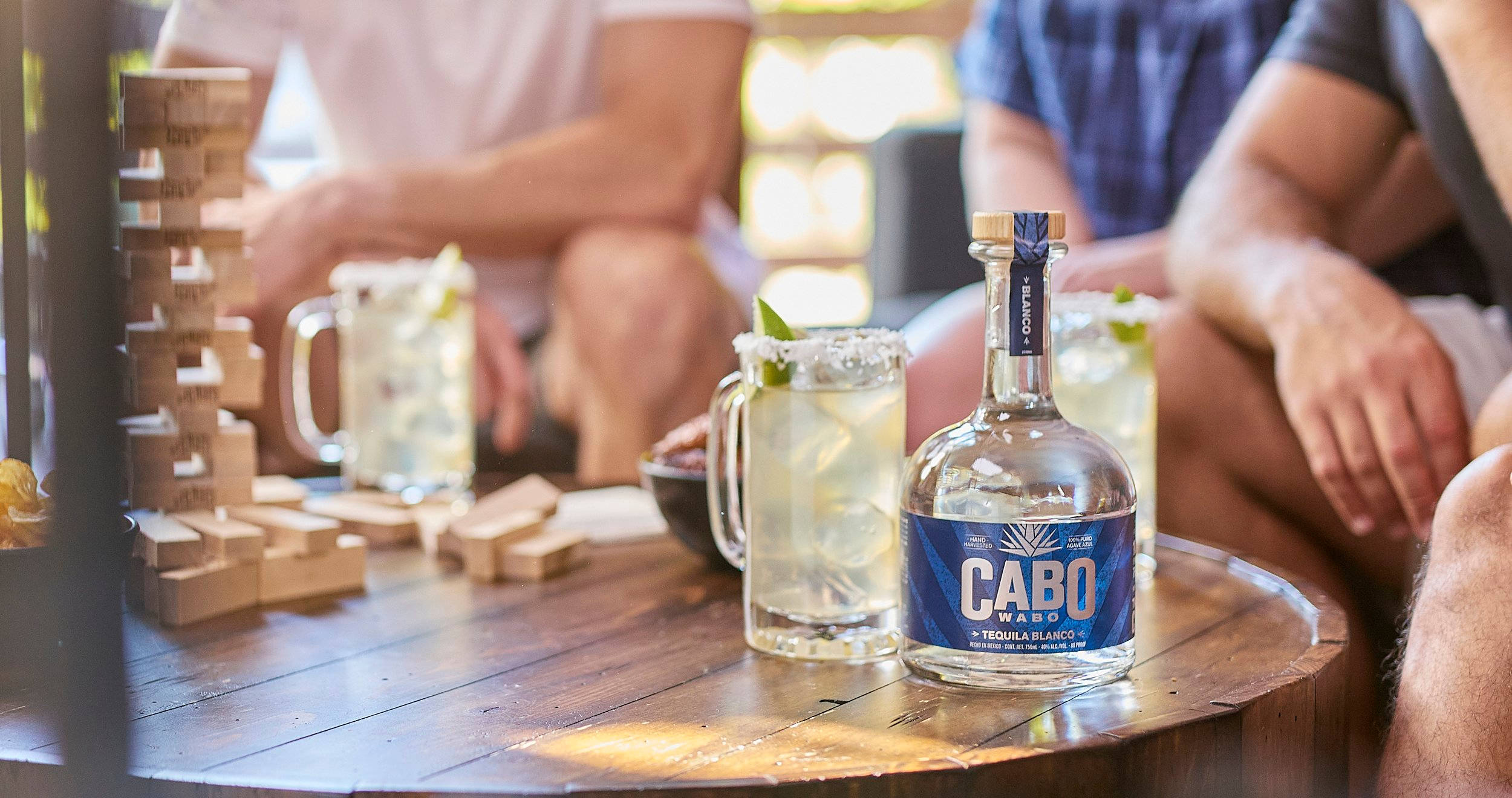 Drink 桌布 壁紙 — 一瓶Cabo Wabo龍舌蘭酒和瑪格麗特酒放在木桌上，旁邊是Jenga遊戲，朋友們在戶外放鬆。