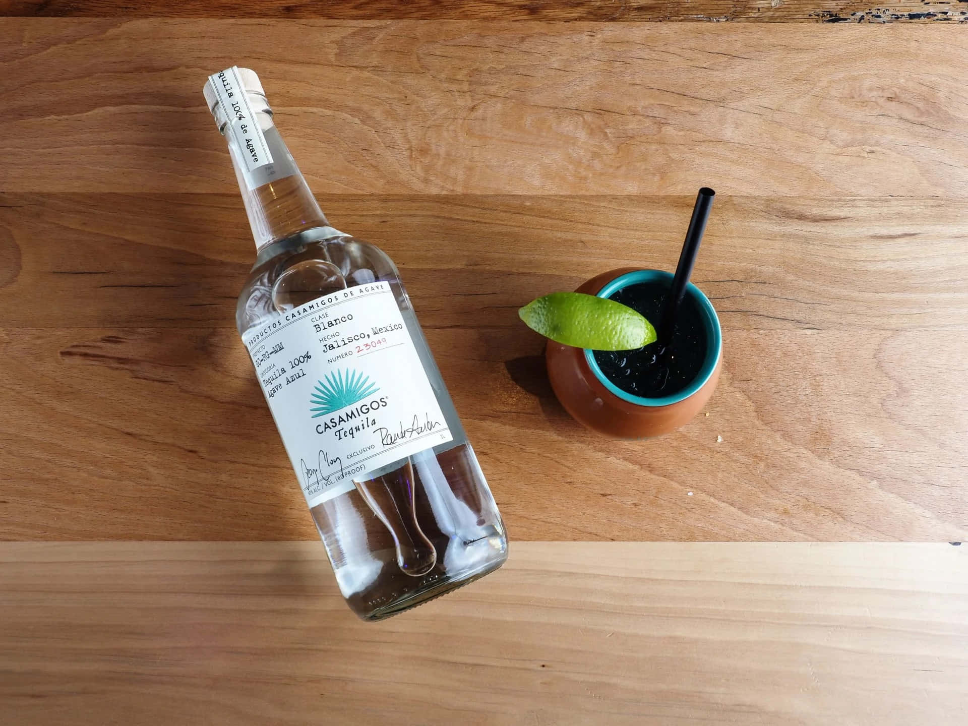 bottle 桌布 壁紙 — 一瓶Casamigos Blanco龍舌蘭酒放在一個鮮藍色邊緣的陶杯旁邊，杯內盛裝著深色雞尾酒，飾以萊姆角，置於溫暖的木質表面上。