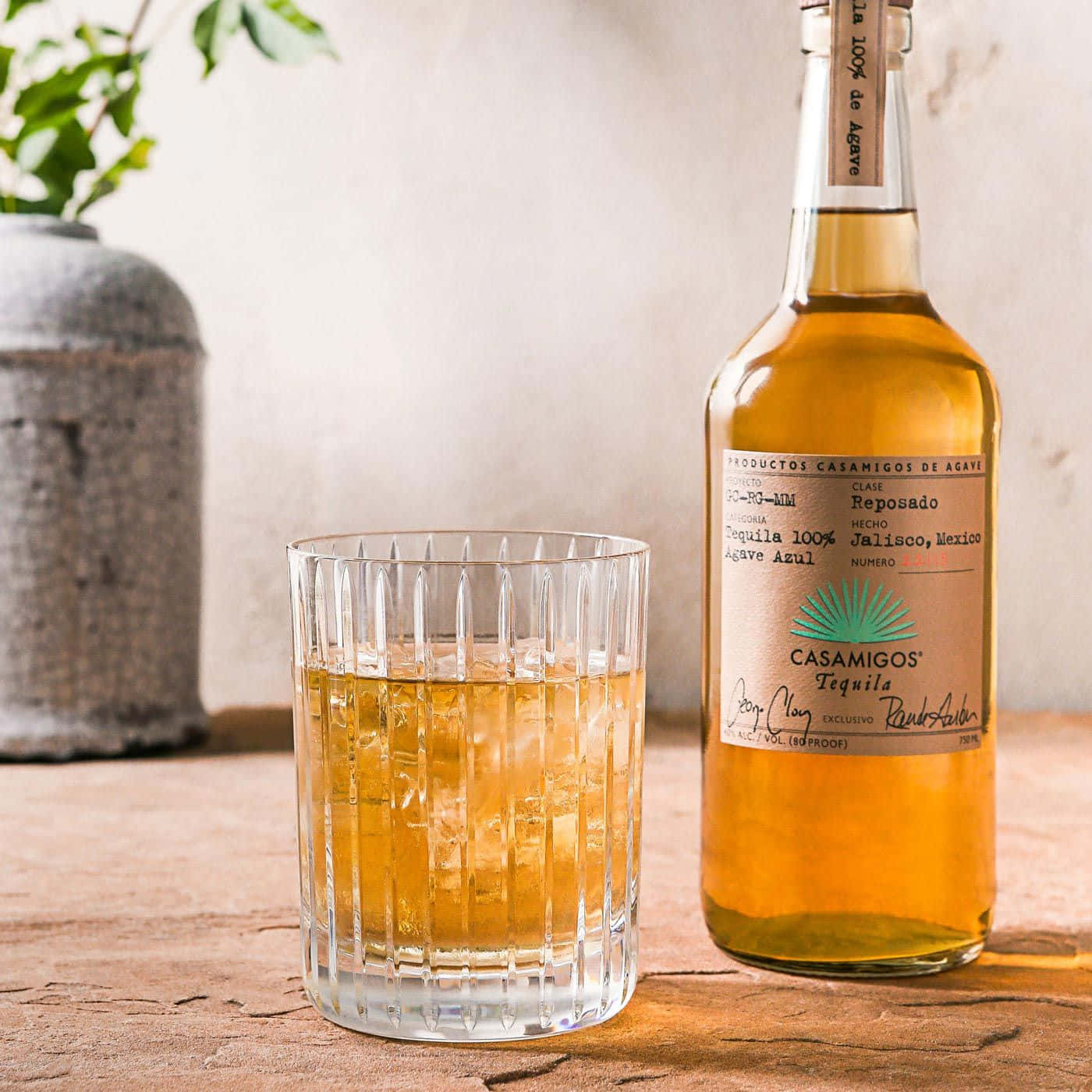 alcohol 桌布 壁紙 — 一瓶Casamigos Reposado龍舌蘭酒和一個裝滿冰塊的玻璃杯放在一個質樸的木桌上，旁邊是一個帶有綠色植物的石花瓶。