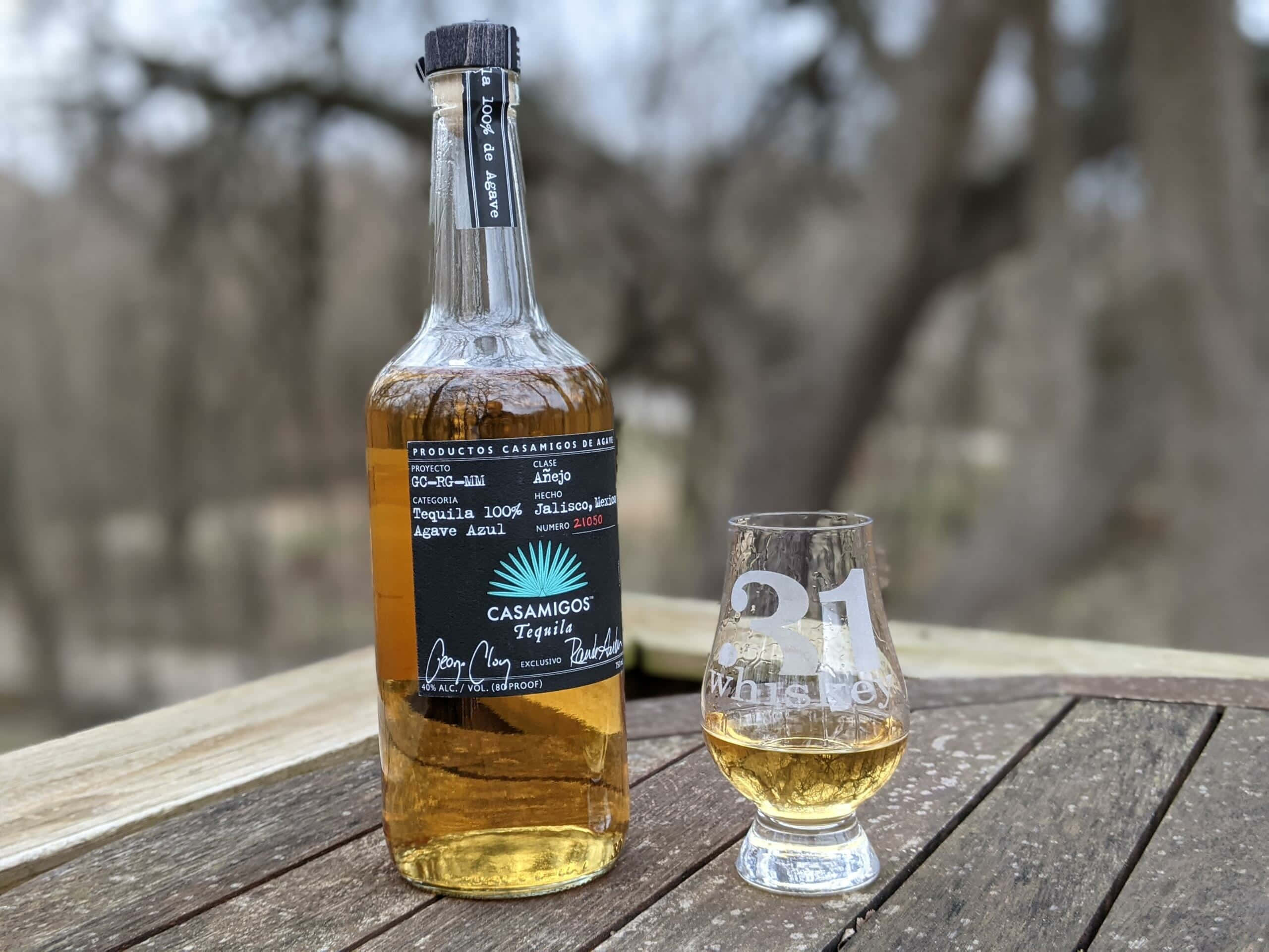 alcohol 桌布 壁紙 — 一瓶Casamigos Tequila和一個裝滿金色液體的品牌玻璃杯放在戶外一張風化的木桌上。